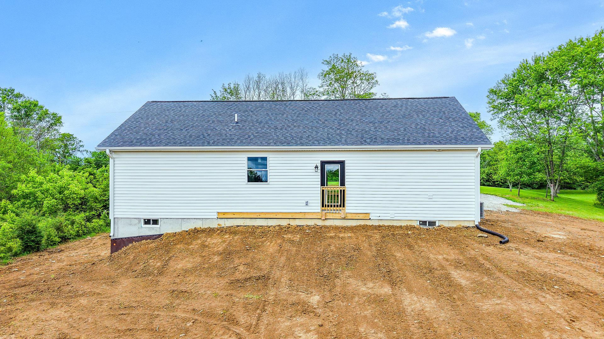 White house with porch and wooden railing, window reflecting trees, wooden gate on window, large dirt hill and patch in front yard, trees and cloudy sky in background.