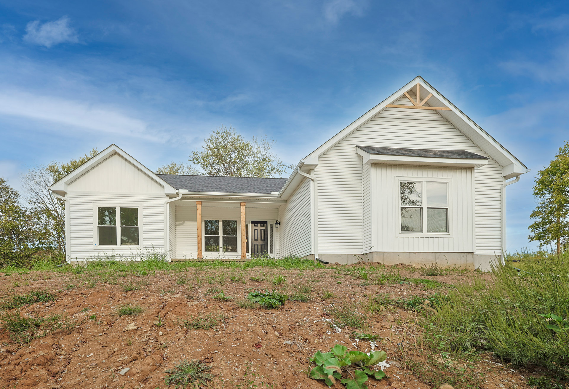 White house with black front door, large windows reflecting nearby trees, green lawn and leafy plants in the yard, dark roof with trees in the background under a partly cloudy sky
