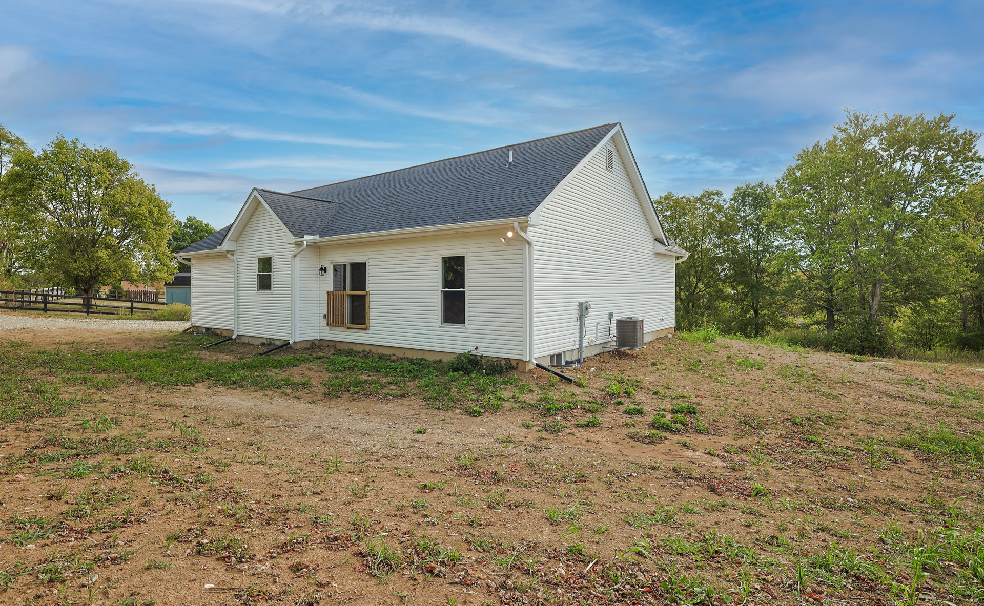 White farmhouse with wooden porch railing, surrounded by dirt field and trees, Robert Frost Farm visible in background under blue sky.