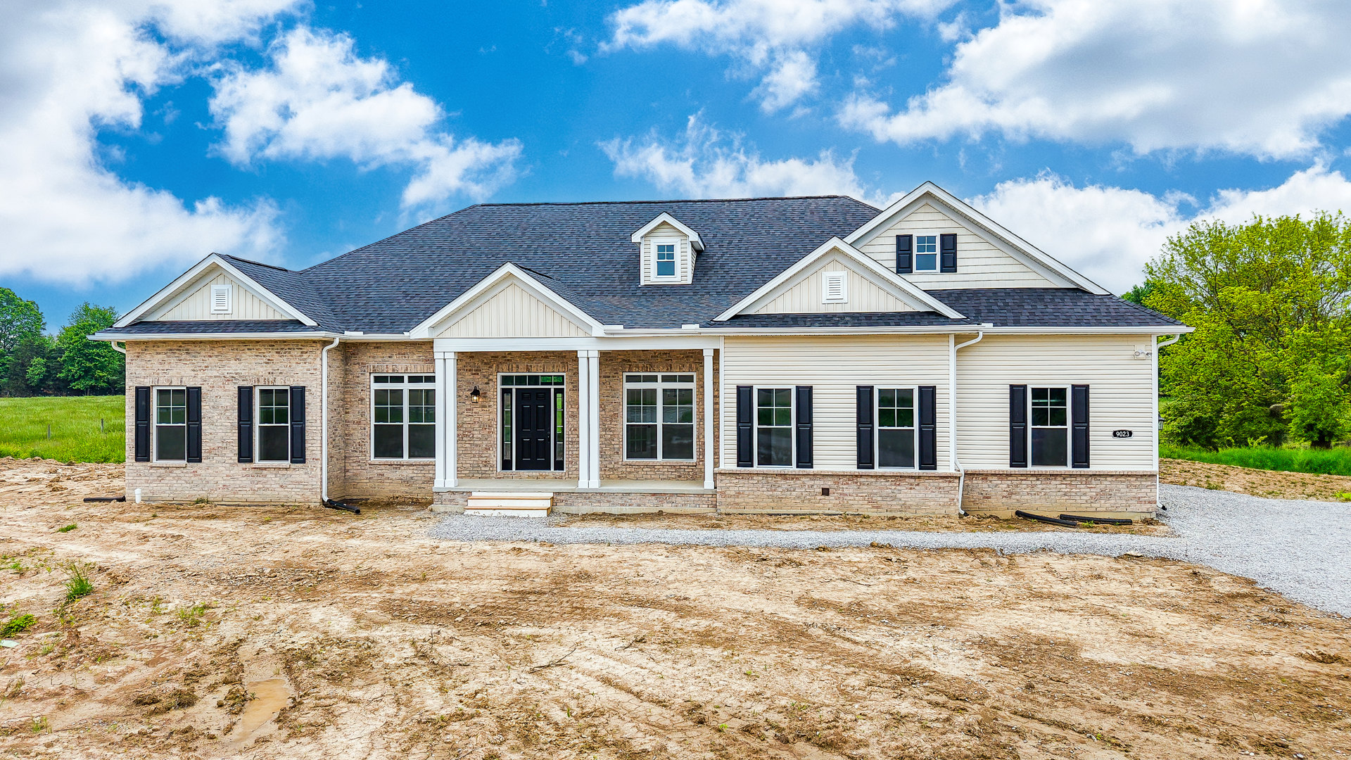 Partially built brick house with black door and glass panes, dormer window on roof, surrounded by dirt field and trees in background