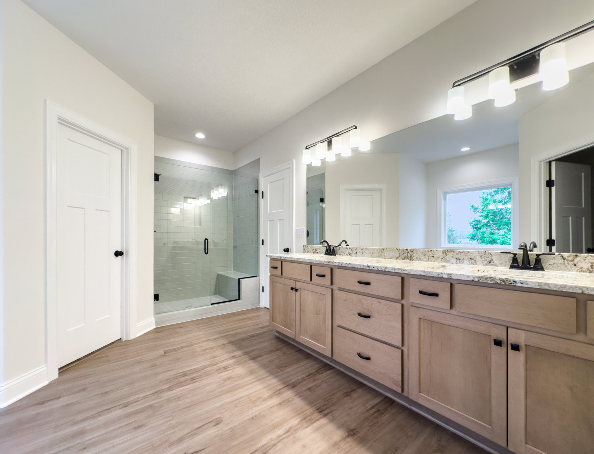 Bathroom with expansive mirror above wide vanity, white door with black knob, window showing trees, glass shower enclosure with bench and shower seat, close-up of wood cabinet