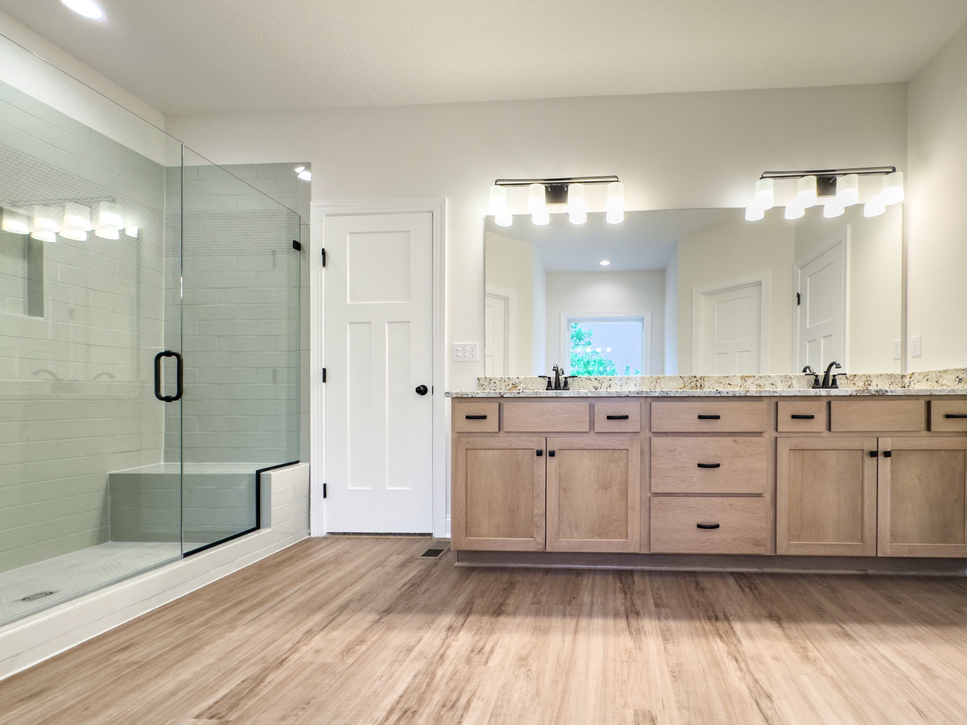 Bathroom with a large wall mirror, glass-enclosed shower, white door with black knob, wood flooring with floor vent, marble kitchen countertop with drawers, close-up of cabinetry