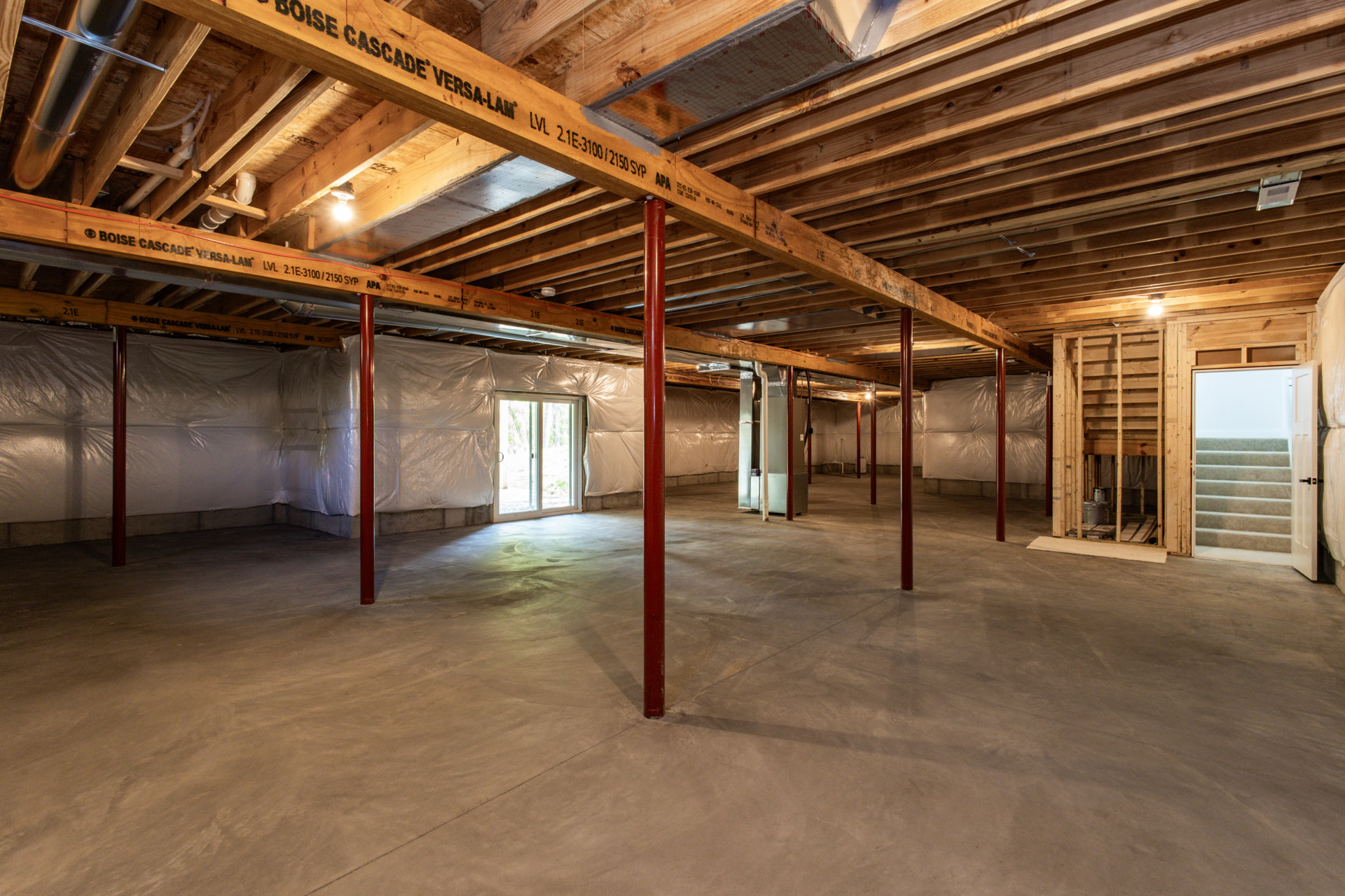 Open-concept room featuring exposed wooden ceiling beams, sliding glass door with white frame, concrete floor with metal support pole, and staircase leading to an adjacent space.