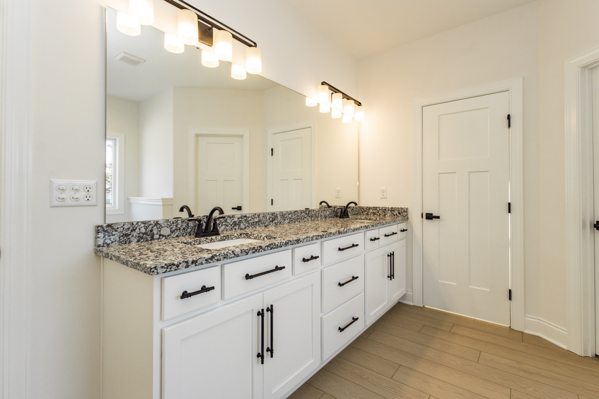Bathroom featuring a marble countertop, large mirror, white door with black handle, wood flooring, white walls, and black hardware accents