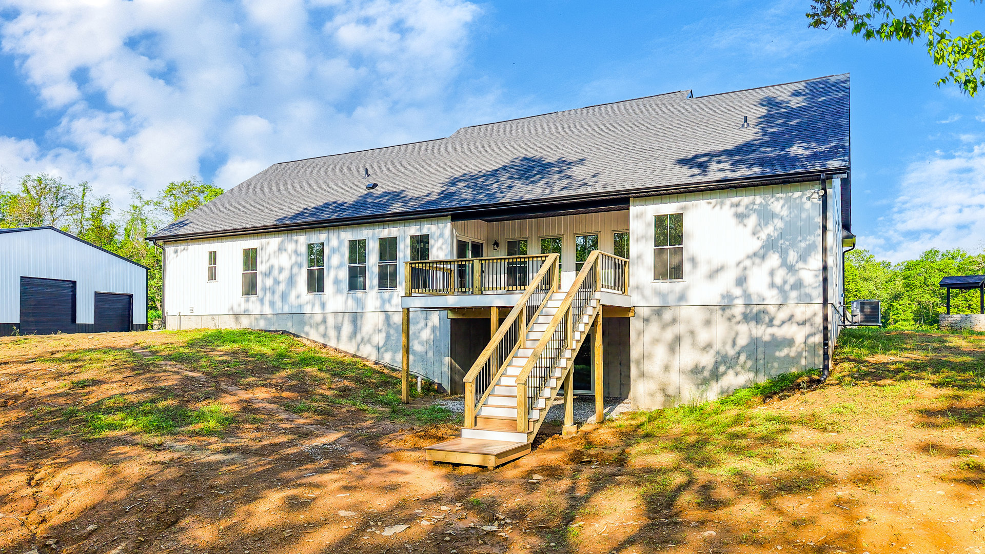 Two-story house with white siding, black front door, wooden deck, and exterior staircase leading to the entrance; surrounded by grass, small trees, and cloudy sky.