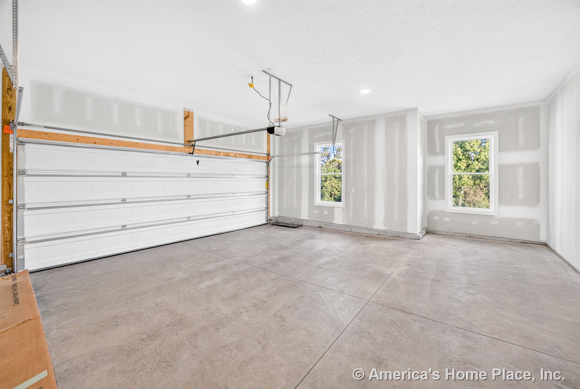 Unfinished drywall garage with concrete slab flooring, large white sectional garage door, ceiling-mounted opener, two double-hung windows, recessed ceiling lights, and minimal