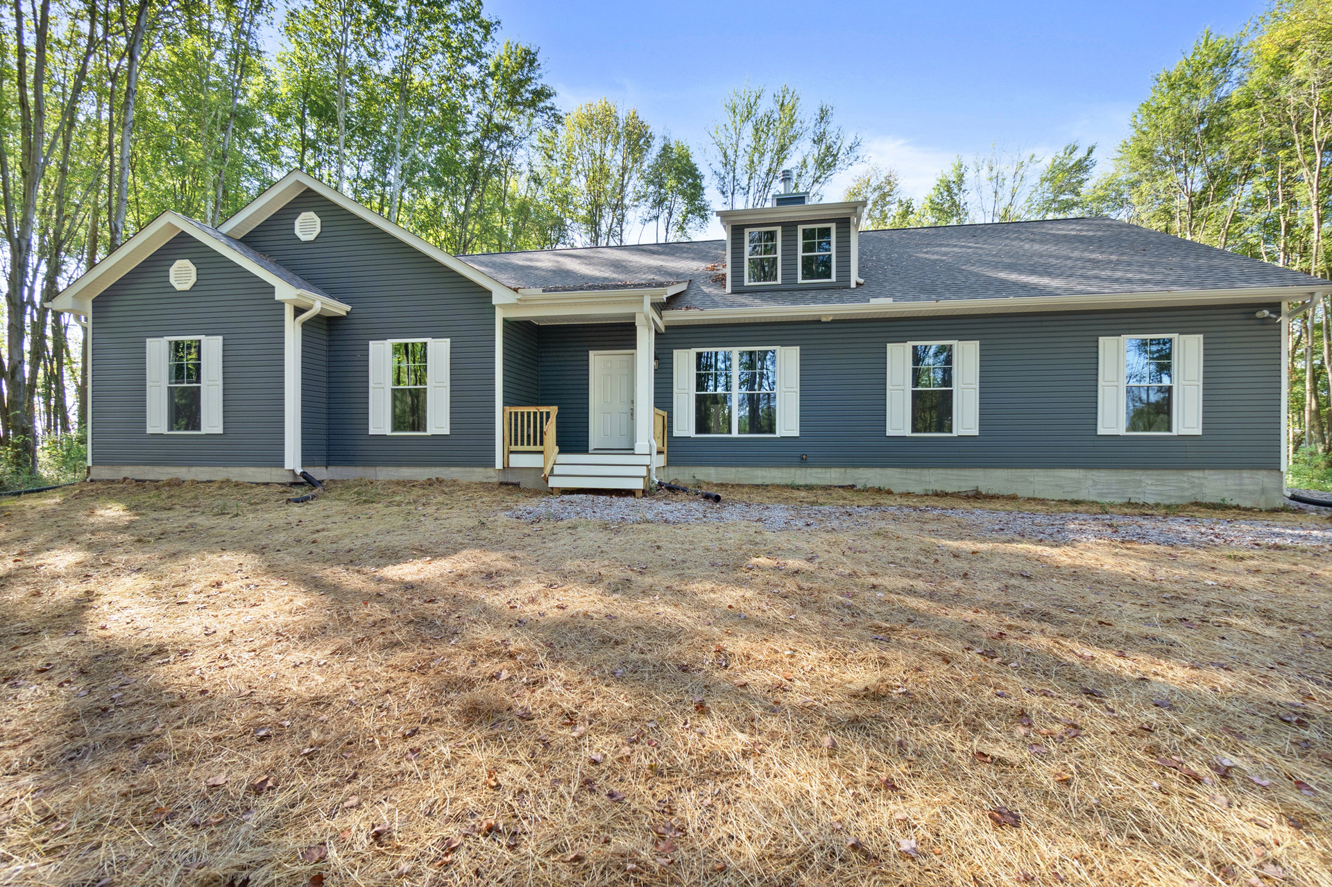 Two-story home with light siding, white shuttered windows, covered porch with wooden railing, grassy yard, trees in background, and small shed to the side