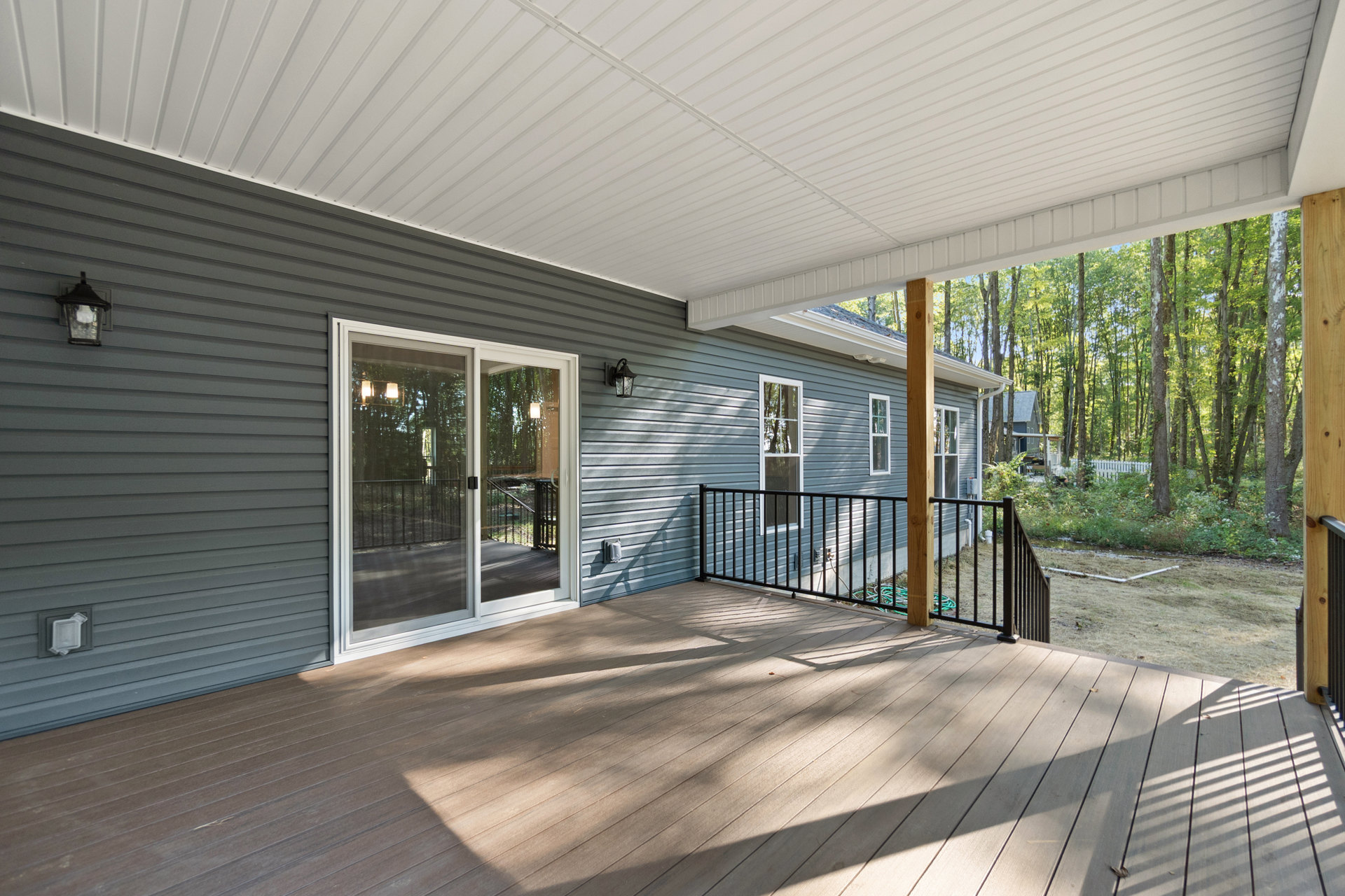 Wood deck with metal railing, covered porch area, sliding glass door, white paneled ceiling, outdoor light fixture, white storage box, partial fence, shaded by nearby trees