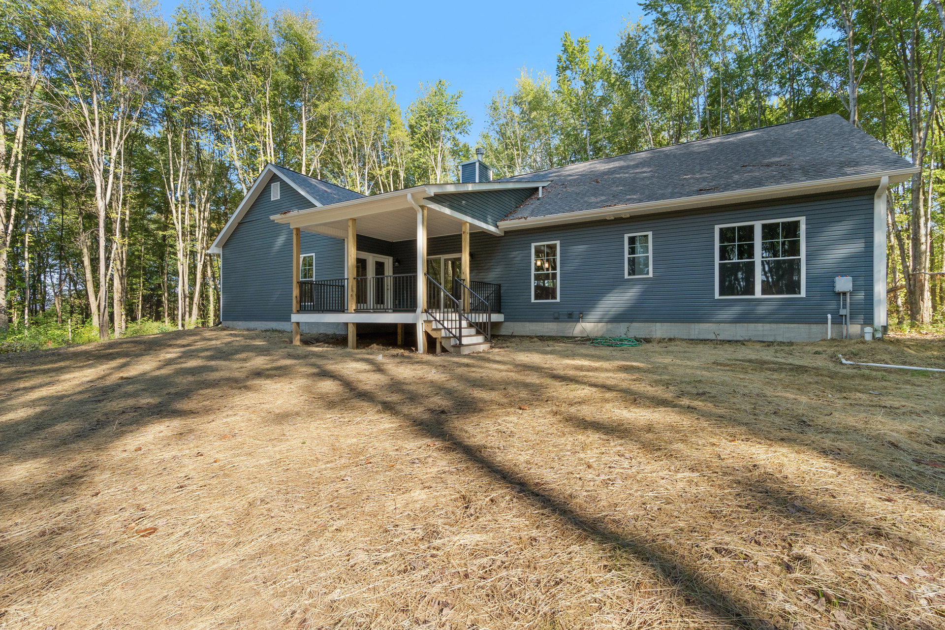 Two-story house with multi-pane windows, white porch railing, front stairs, grassy yard, and wooden fence; mature trees and blue sky in background
