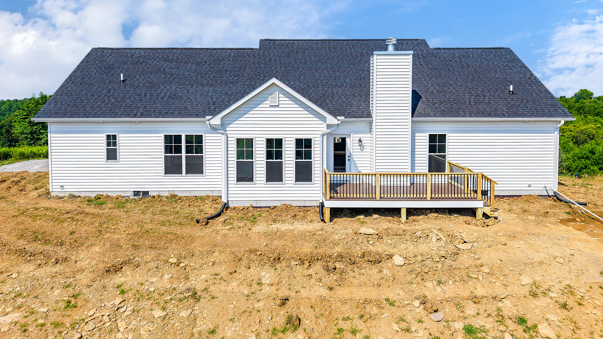 White siding house with wooden deck and railing, fenced yard, large windows with white frames, cloudy sky overhead, green plants along property edge