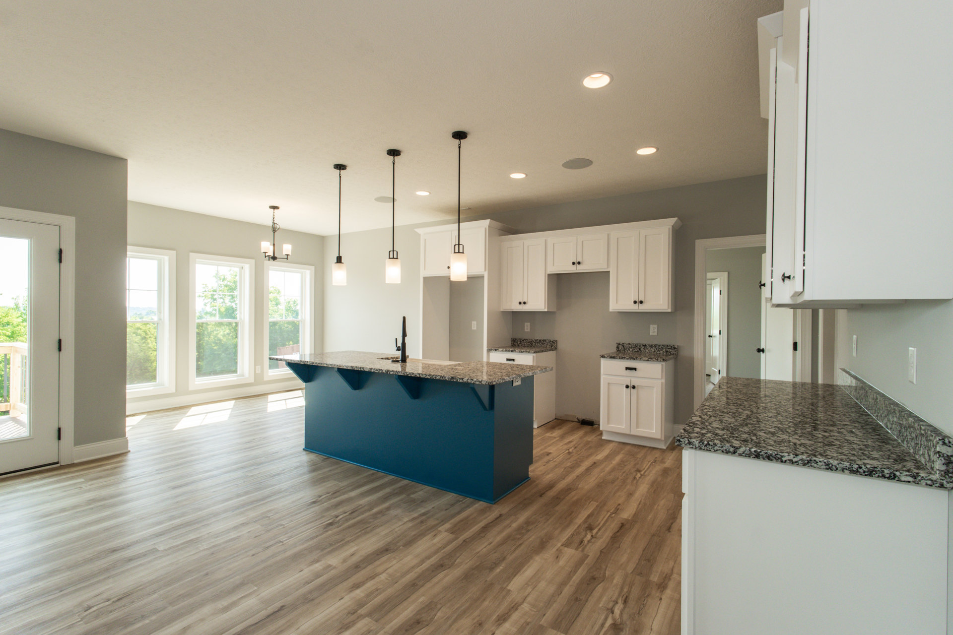 Blue kitchen island with marble countertop, white shaker cabinets, stainless steel sink, tile backsplash, pendant lighting, large window overlooking trees, hardwood flooring