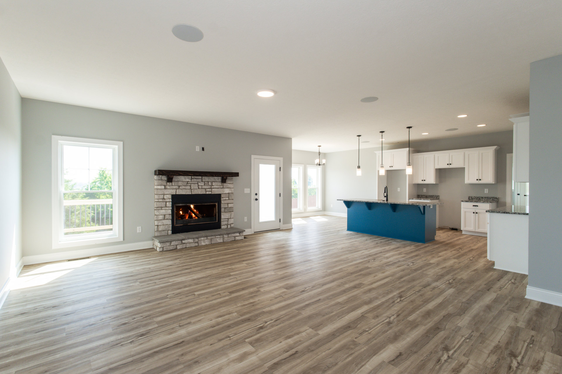 Spacious living area with hardwood floors, stone fireplace with active fire, built-in bar featuring blue countertop, large window overlooking trees, and white door with glass panel