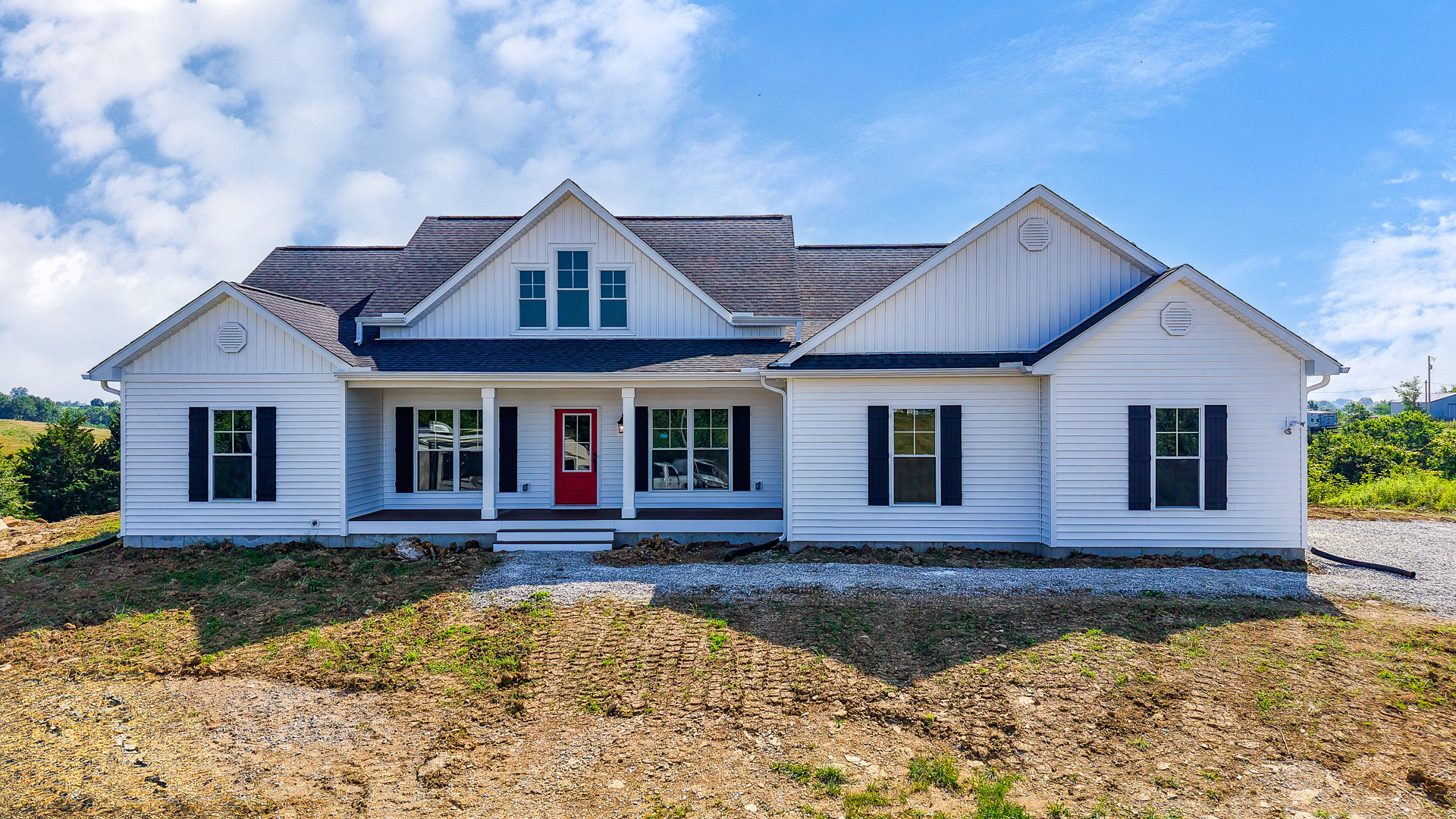 White siding house with red front door, white framed windows, and shutters, set against a cloudy sky with plants near the porch.
