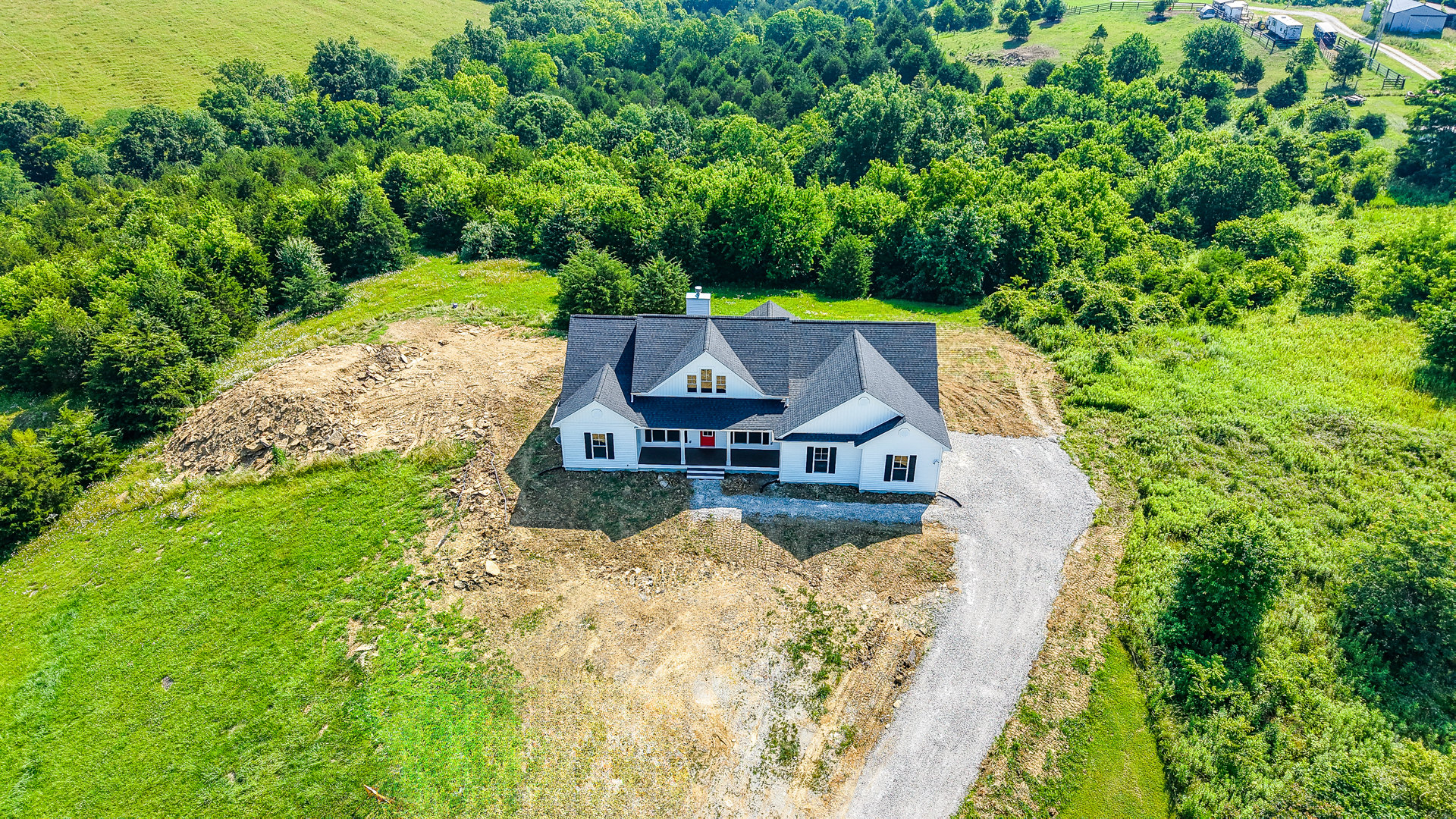 Two-story house with black roof and red door, set on a grassy hill surrounded by mature trees, gravel driveway with drainage pipe in foreground
