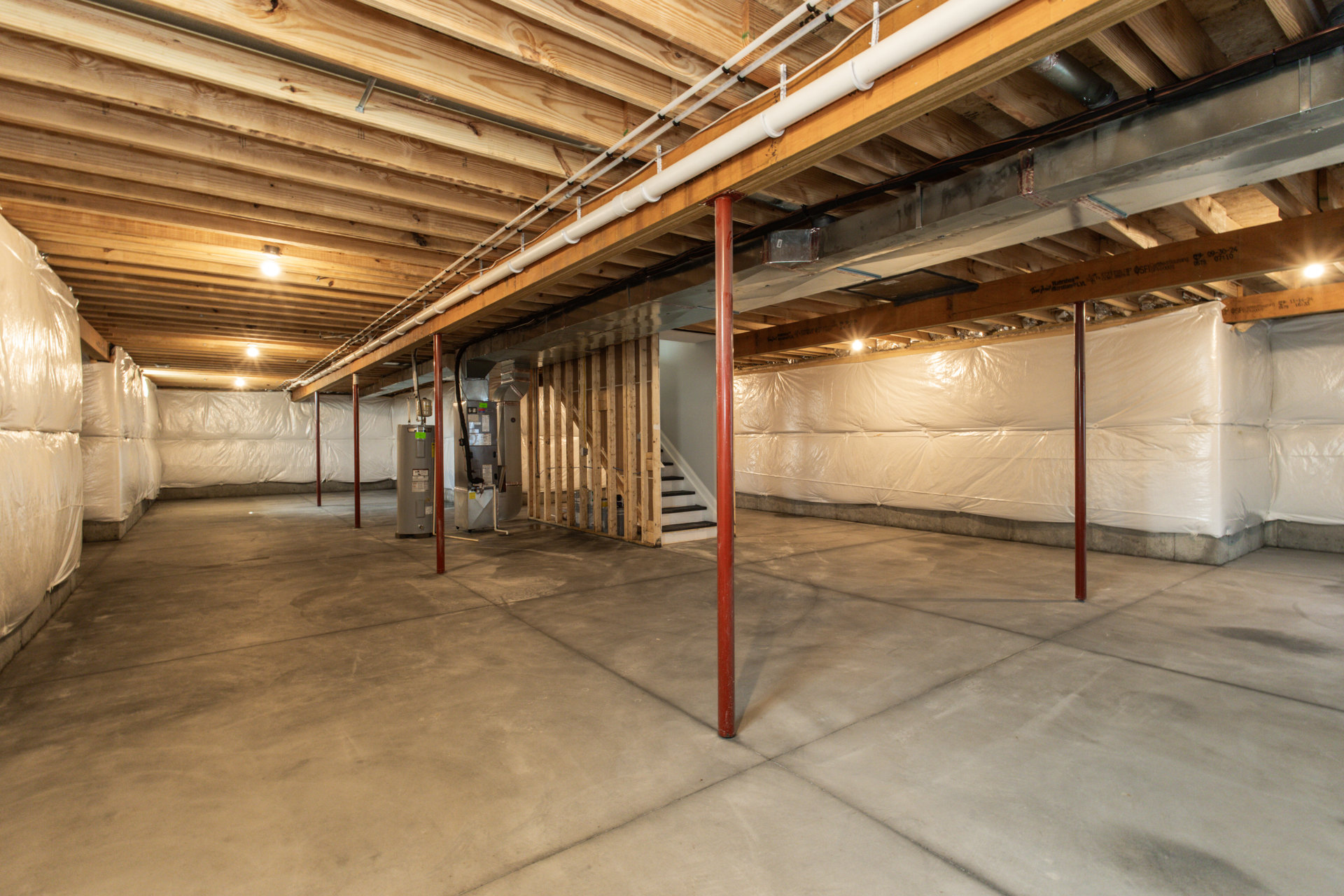 Basement with unfinished wooden ceiling, red steel support pole, white pipe along wall, and plastic-wrapped storage container near short staircase.