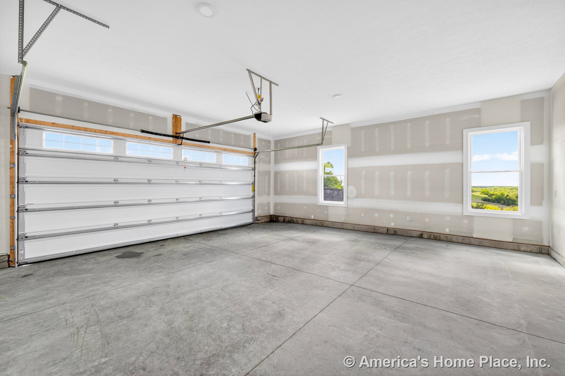 Unfinished drywall garage with concrete flooring, large sectional overhead door featuring upper windows, two side windows with trim, and ceiling-mounted garage door opener.