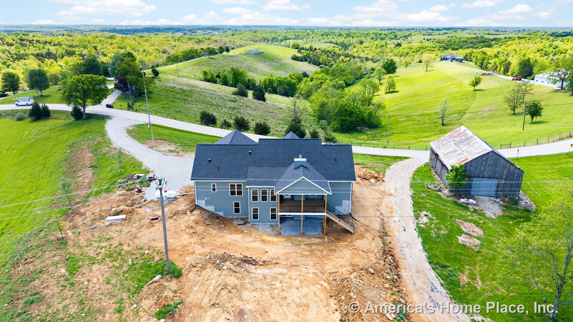 Gray siding exterior with multiple rectangular windows, dark shingle roof, covered rear porch supported by wood posts, walkout basement, and gravel driveway set against a rural