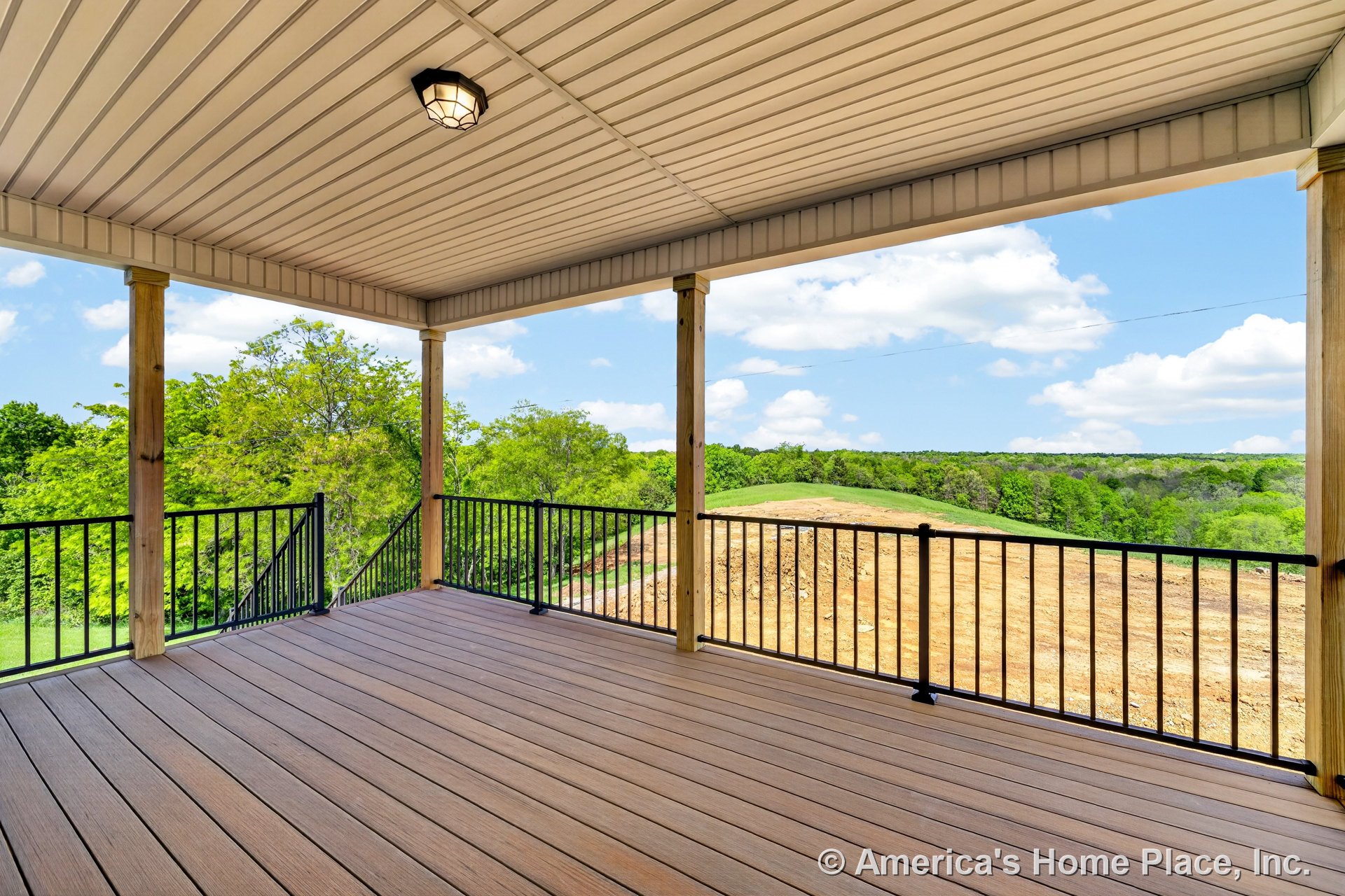 Rear deck with composite flooring, covered ceiling featuring built-in recessed lighting, black metal railings, and wood support columns overlooking an open outdoor area.