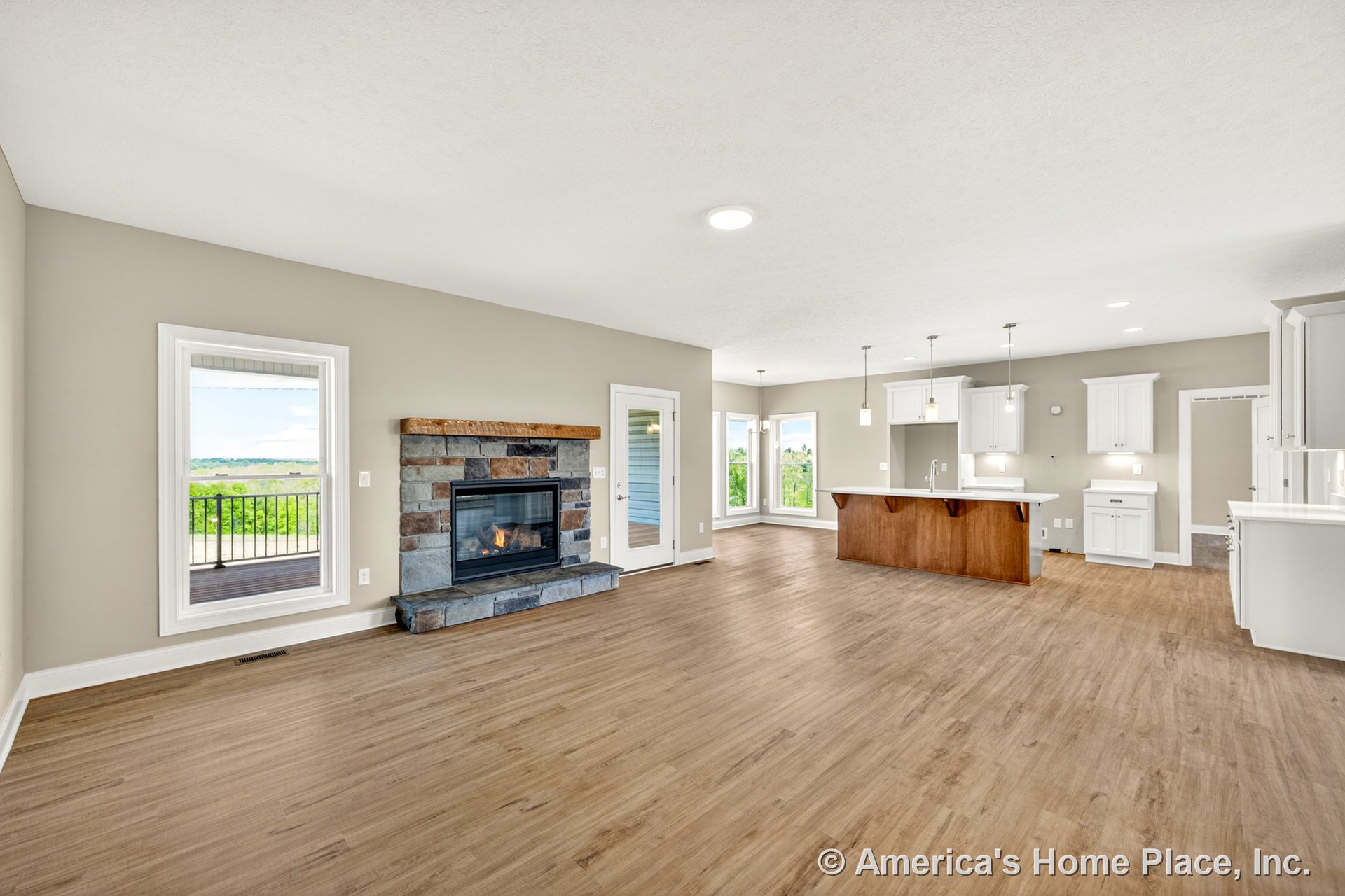 Stone fireplace with wood mantel beside large windows with white trim, open living area with wood-look plank flooring, kitchen island with wood finish, white upper and lower