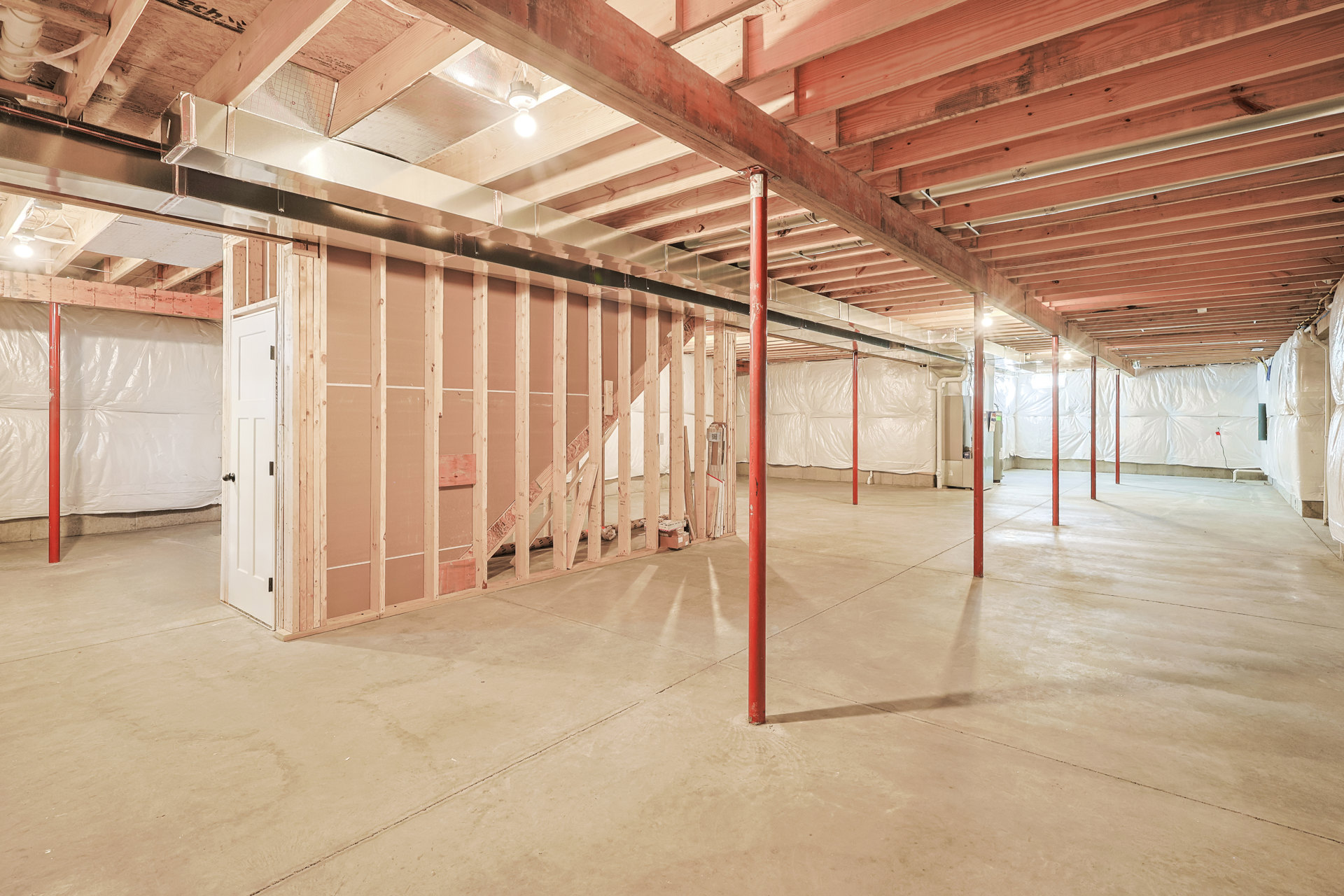 Basement room with exposed red steel support poles, white paneled door with silver doorknob, unfinished wooden ceiling beams, concrete floor, and visible light bulb fixture