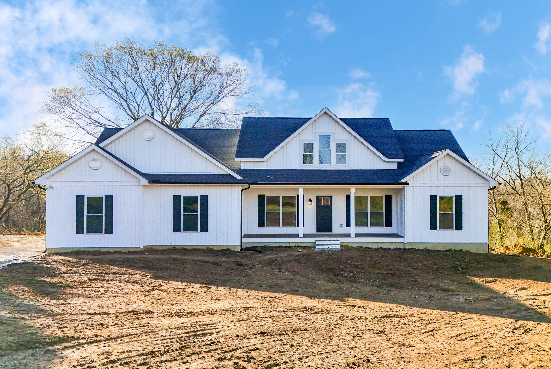 White house with black roof and door, three windows with white frames, surrounded by large dirt field, white fence along property edge, cloudy sky overhead.