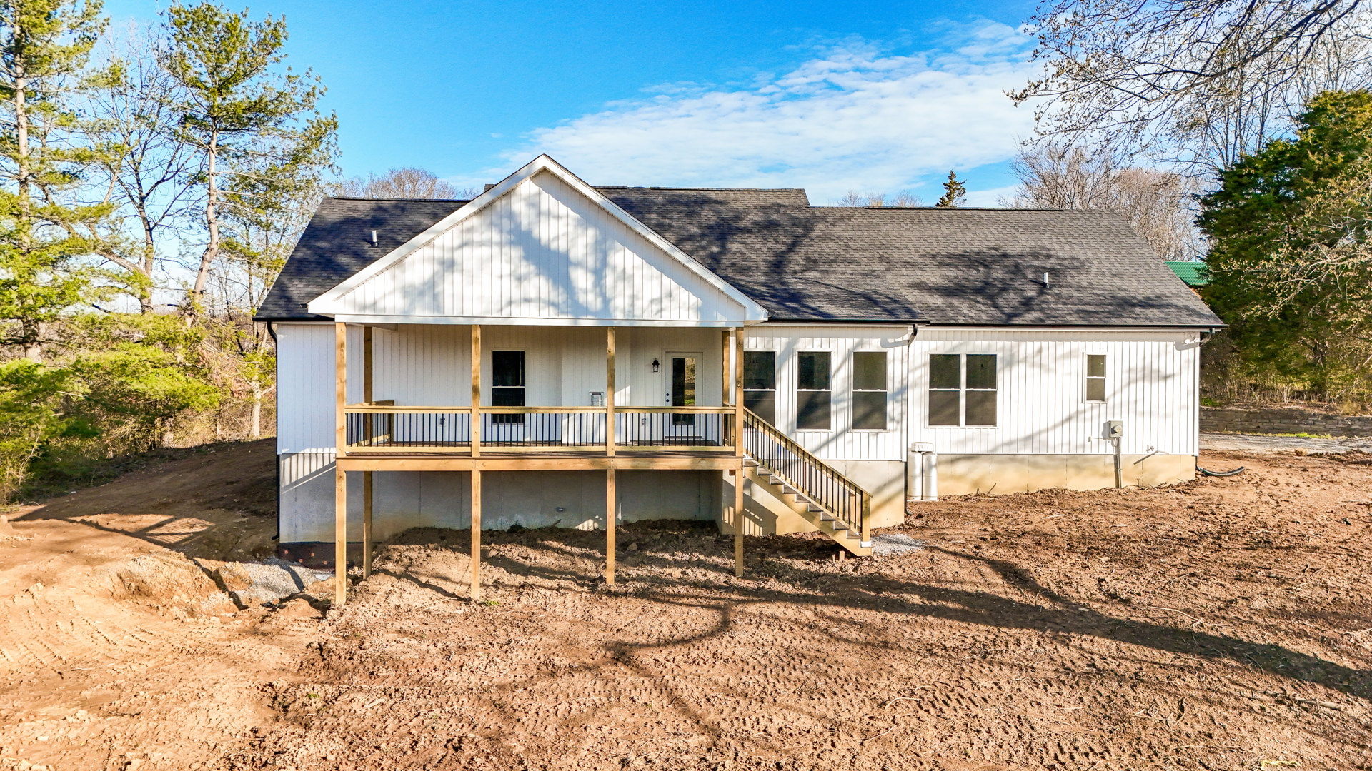 Two-story home with white siding, covered front porch, wooden deck and railing, white-framed windows, fenced yard, and landscaped greenery under a partly cloudy sky