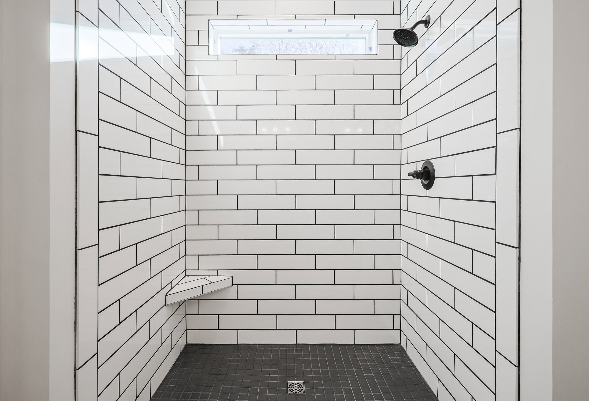 White tile shower with black grout lines, window centered above a blue accent, black shower head and pole, floor drain, modern farmhouse style.
