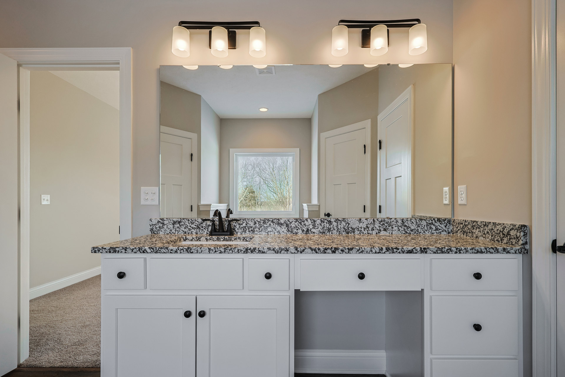 Bathroom with marble countertop, undermount sink, large wall mirror, white cabinetry with drawers, black hardware, white door with black handle, window overlooking trees, and