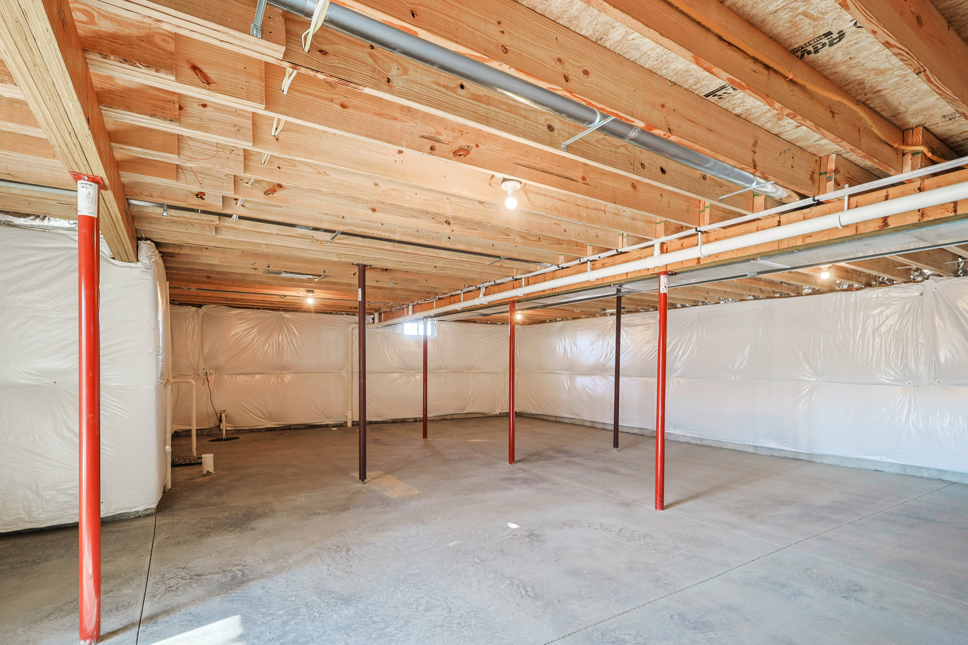 Basement room with exposed concrete floor, white sheet covering ceiling, visible pipes, red poles, and light bulb fixture