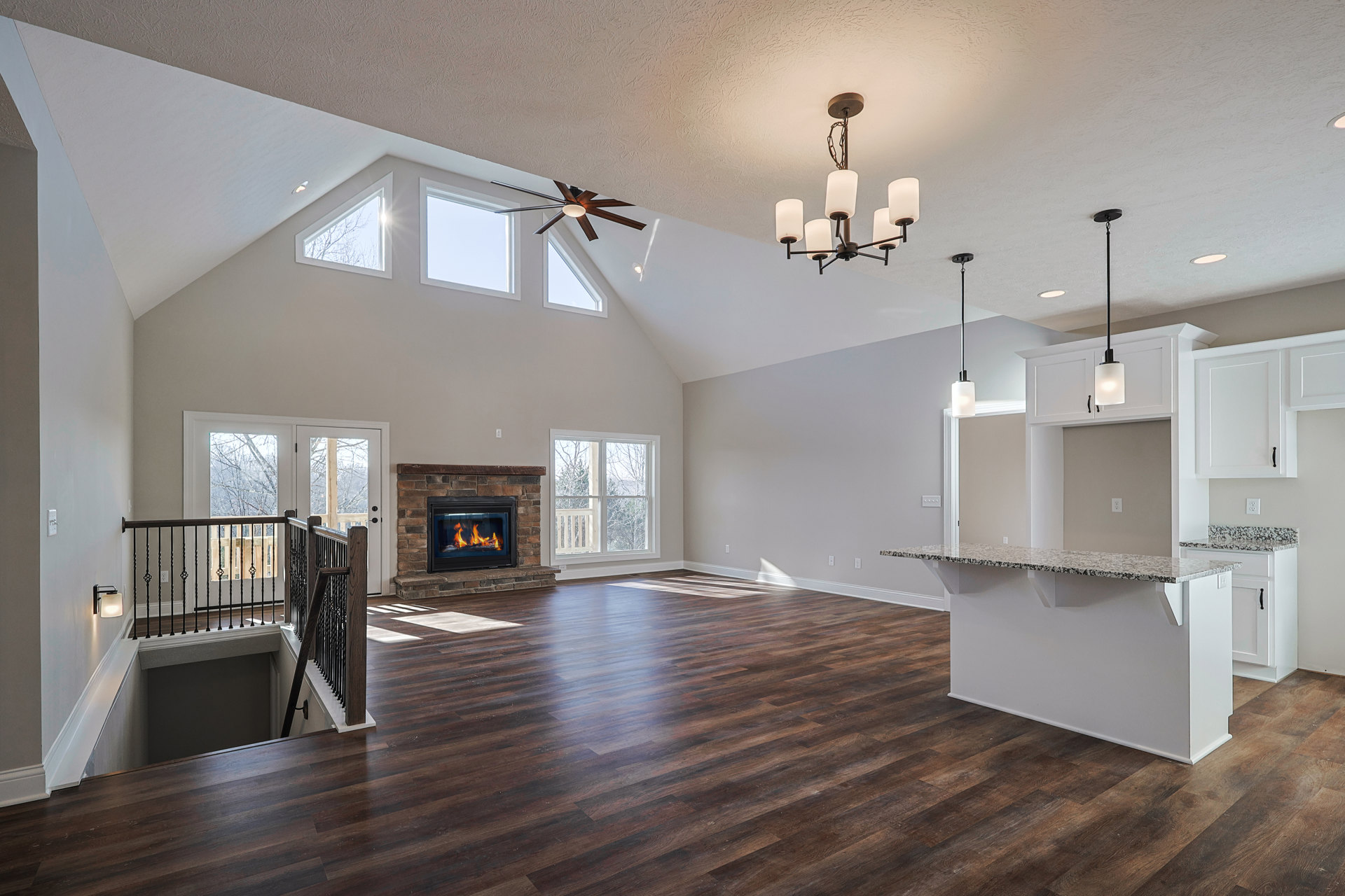 Spacious living room with hardwood floors, white plaster walls, ceiling fan, active fireplace, and large window overlooking trees