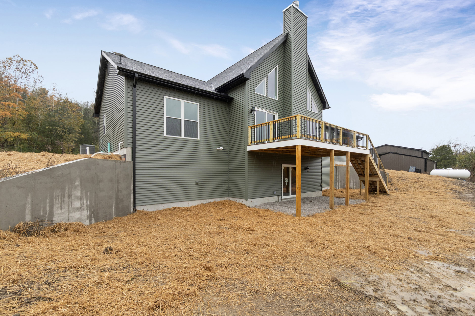 Two-story house with wood deck and railing, fenced yard, large windows, straw scattered on concrete patio, trees and cloudy sky in background