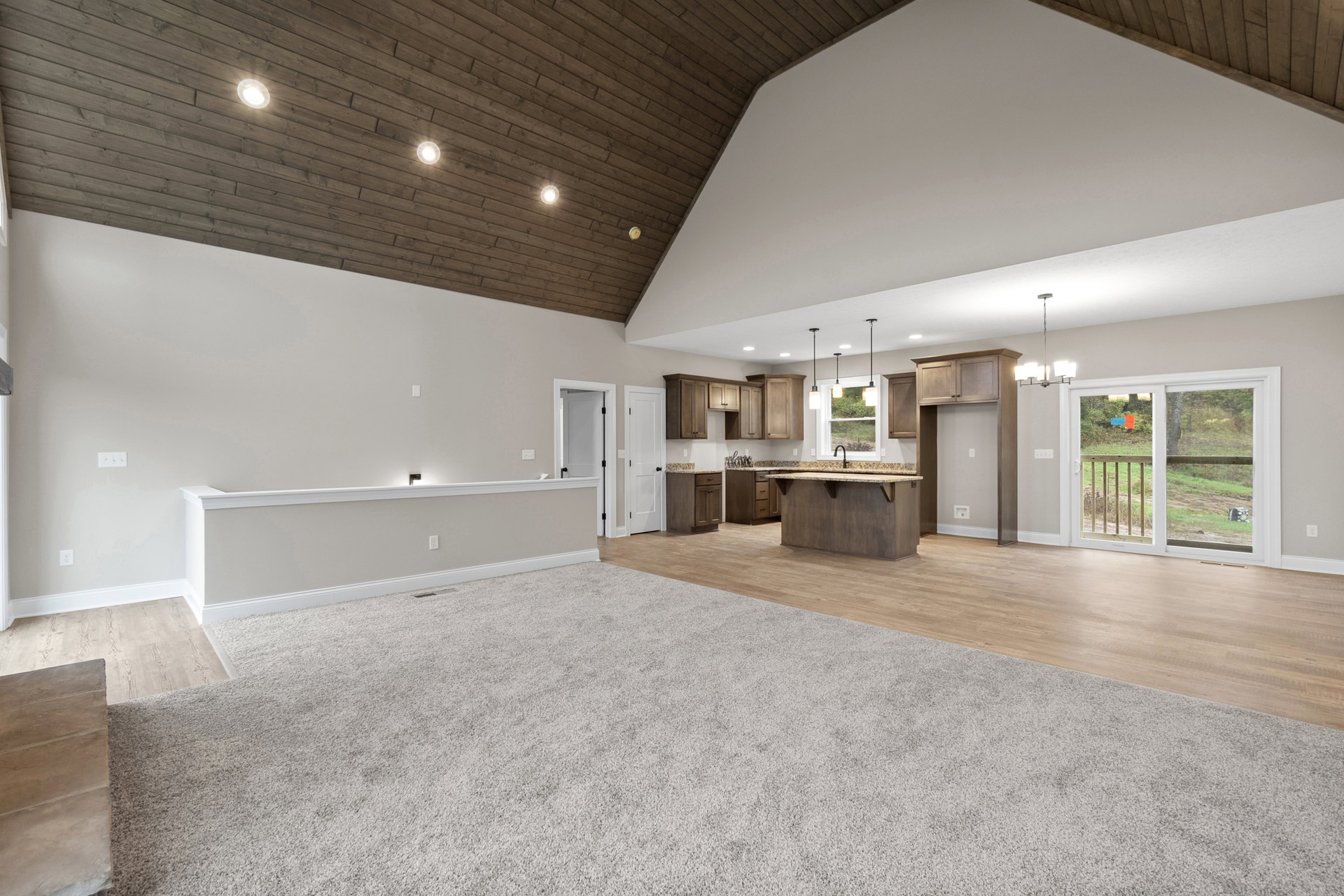 Open-concept kitchen and living area with high ceiling, marbled countertop, wood cabinetry, white carpet and wood flooring, sliding glass door leading to wooden deck with railing.