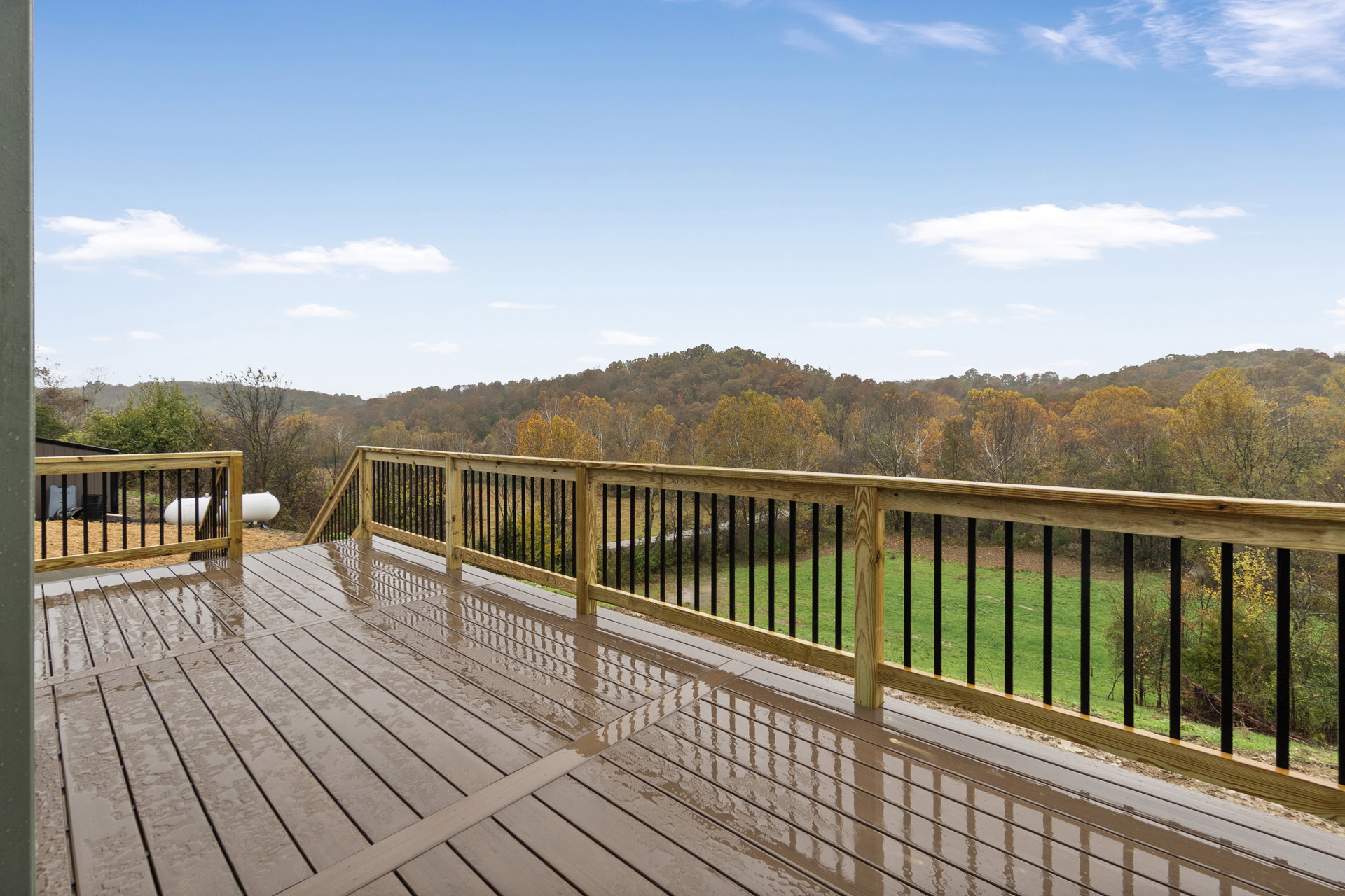 Wooden deck with railing overlooking grassy yard and cluster of trees, fence visible in background under partly cloudy sky