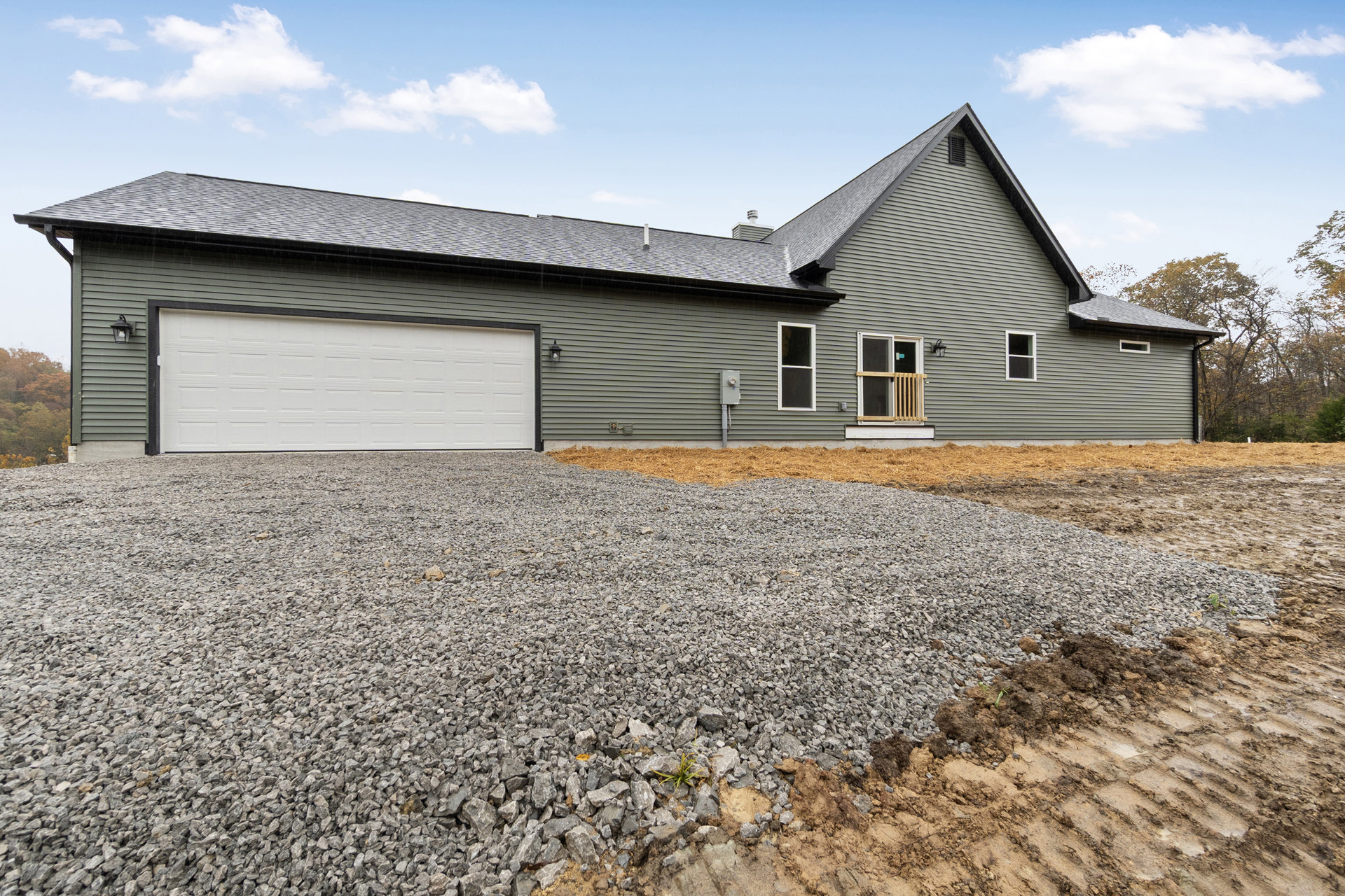Gravel driveway leading to white garage door with black trim, adjacent window with white frame and wooden railing, surrounded by trees and plants under cloudy sky