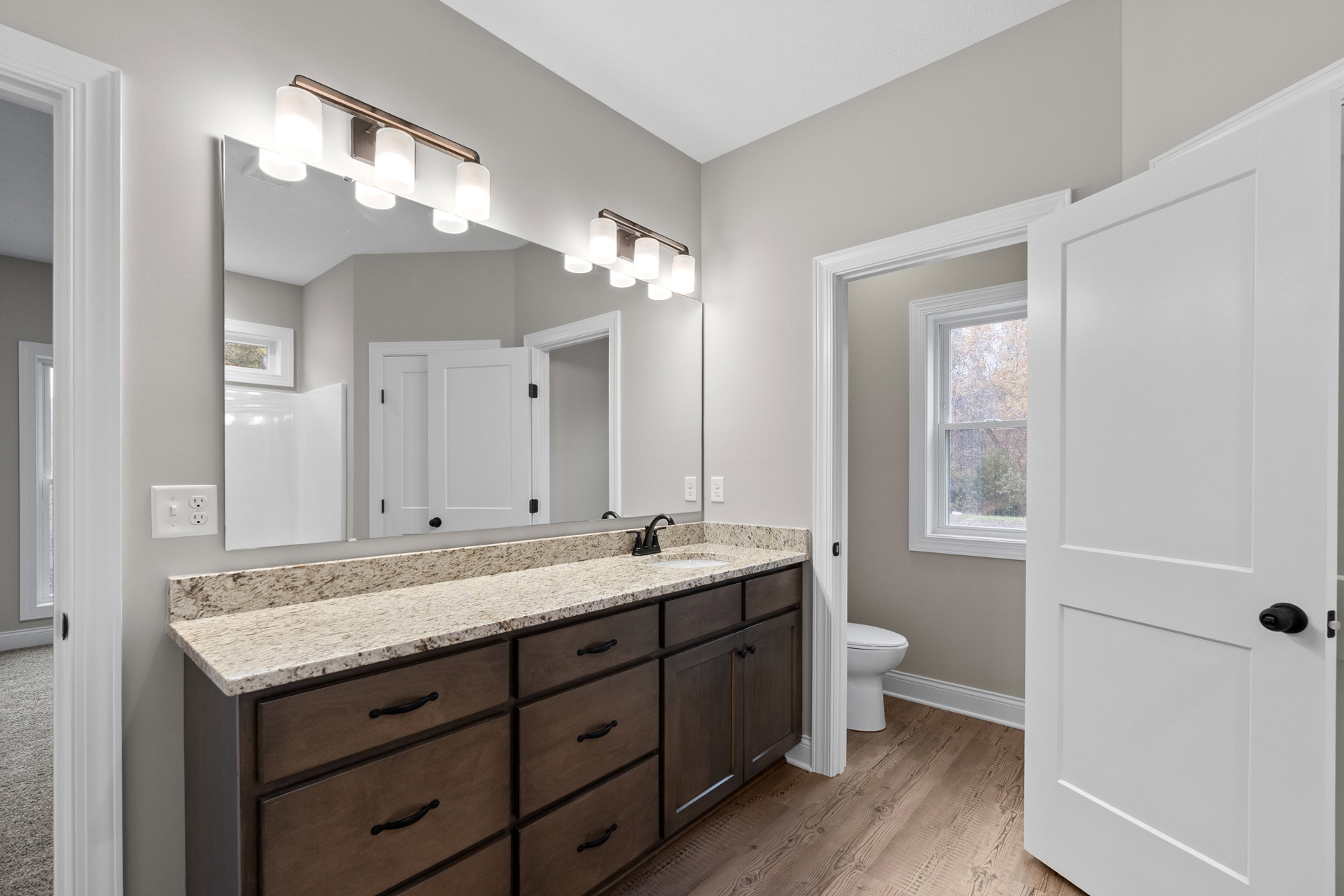 Bathroom with tiled walls, large framed mirror above a stone countertop, modern sink and faucet, white cabinetry with drawers, toilet, window overlooking trees, and a light switch