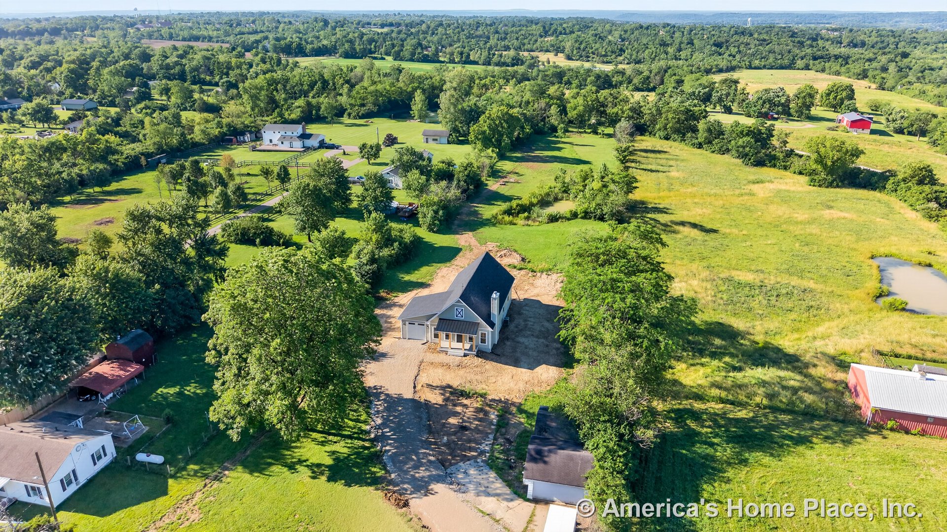 Covered front porch with columns, attached two-car garage, prominent gable roof with chimney, large front-facing windows, concrete driveway, and landscaped rural surroundings with