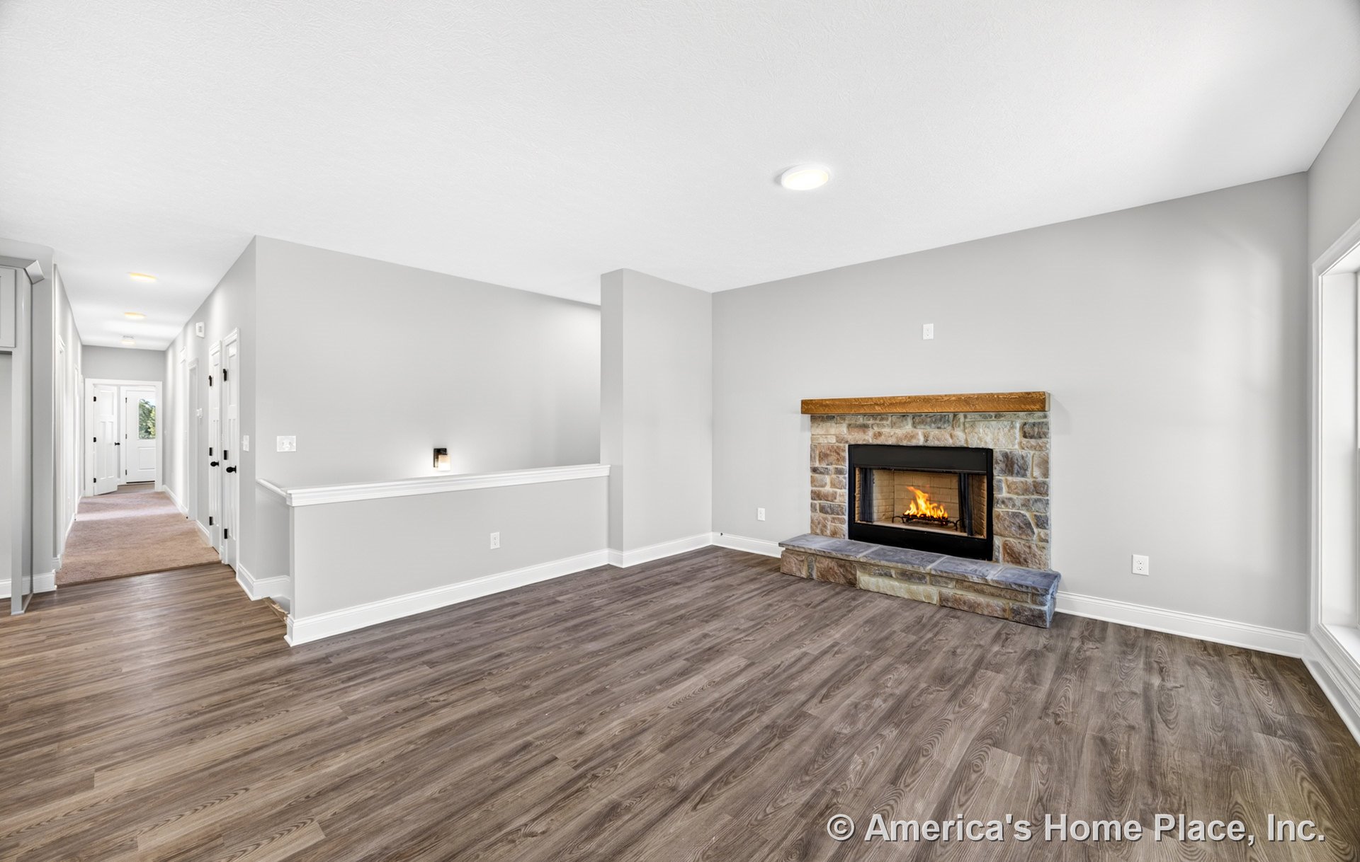 Stone fireplace with wood mantel, wood-look vinyl plank flooring, neutral gray walls with white trim and baseboards, recessed ceiling lighting, open hallway with multiple doors.