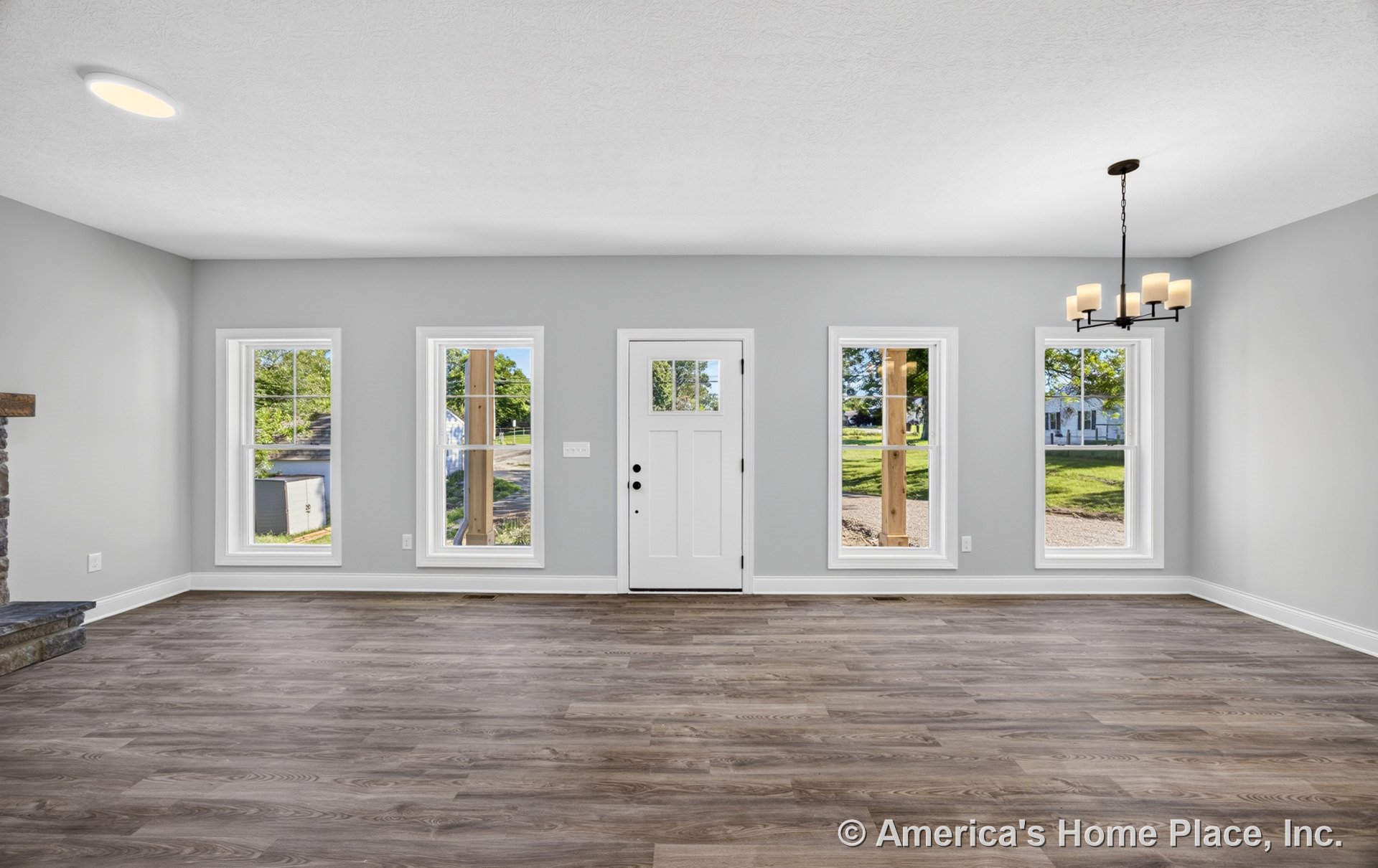 Spacious living area with four large divided-light windows, white front door, wood-look vinyl plank flooring, gray painted walls, white trim and baseboards, modern chandelier, and