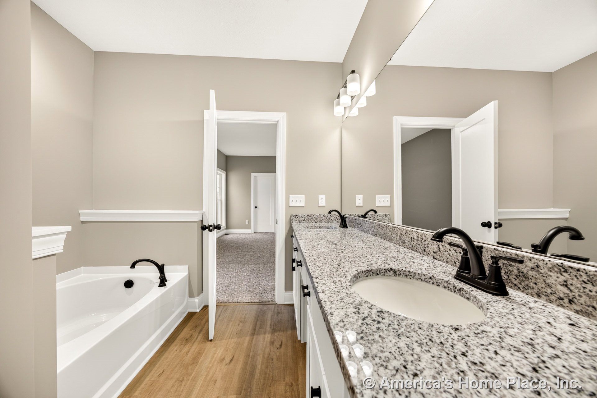 Granite double vanity with white cabinetry and matte black faucets, large wall mirror above, built-in soaking tub beside, wood-look flooring, neutral wall color, and trim molding.