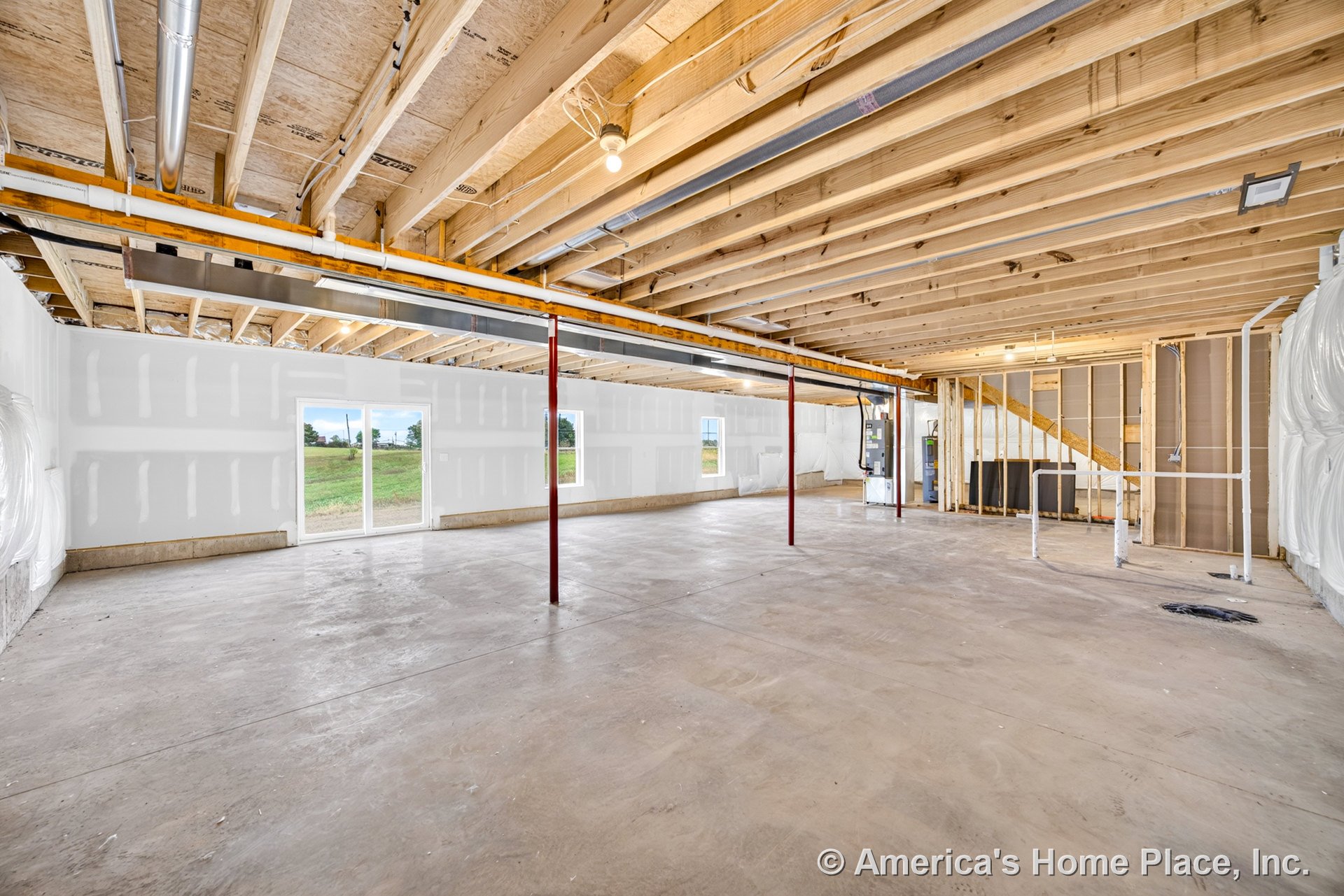 Unfinished basement with exposed wood ceiling joists, concrete slab floor, drywall walls with visible seams, support columns, open staircase framing, utility pipes along one wall