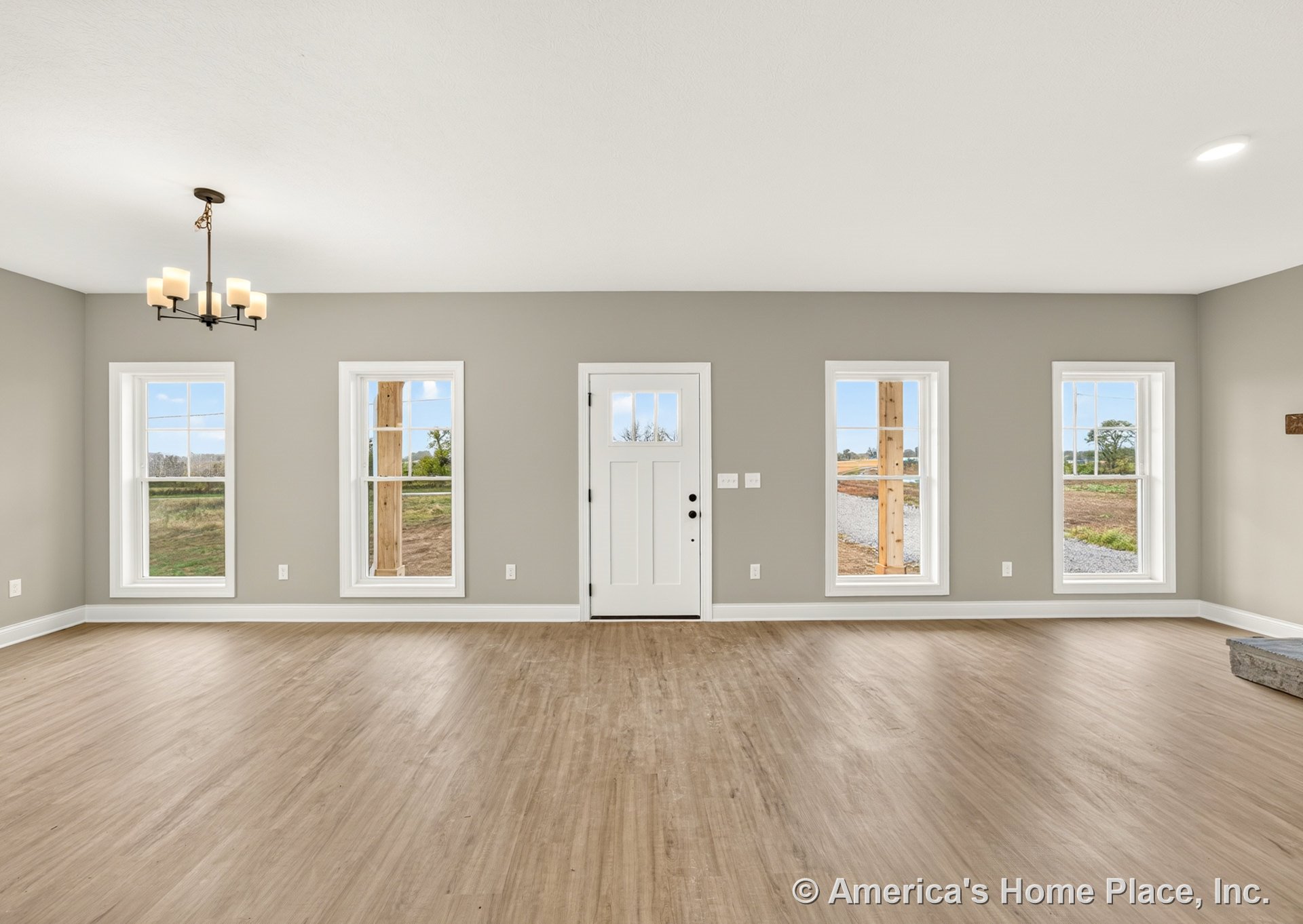 Living area with light wood plank flooring, gray painted walls, five tall windows with white trim, white paneled front door, white baseboards, and a modern chandelier centered on