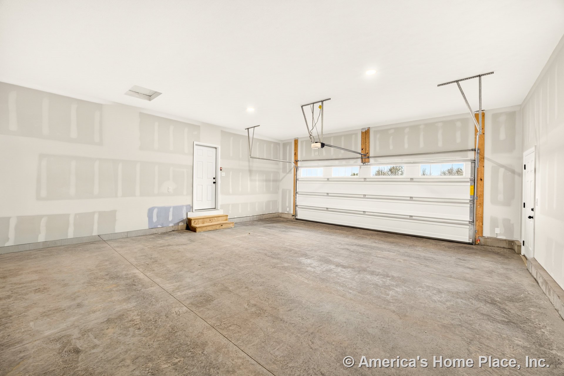 Unfinished garage with exposed drywall walls, concrete slab floor, large sectional overhead door featuring multiple windows, ceiling-mounted light fixtures, and wood steps leading