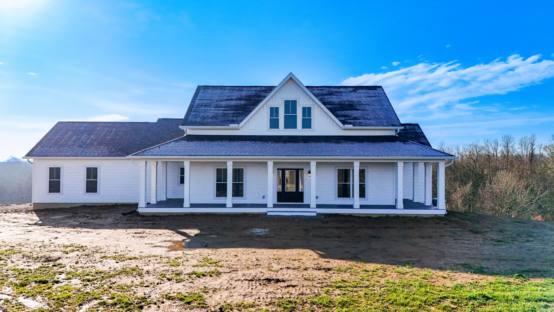 White house with three windows, black front door, white columns, and white fenced yard with patches of grass and dirt under a cloudy sky
