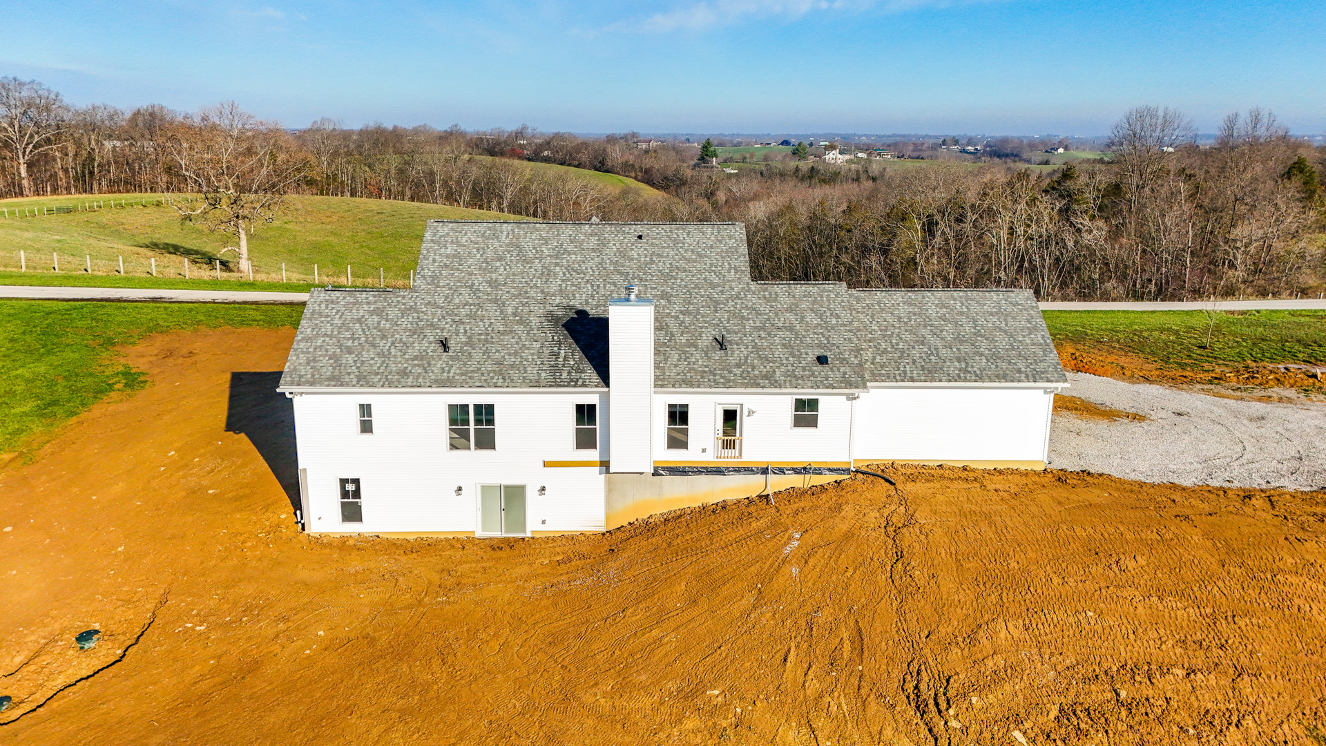 Modern white house with glass-paneled door and horizontal accents, situated on grassy field near dirt hill with tire tracks, surrounded by trees under cloudy sky