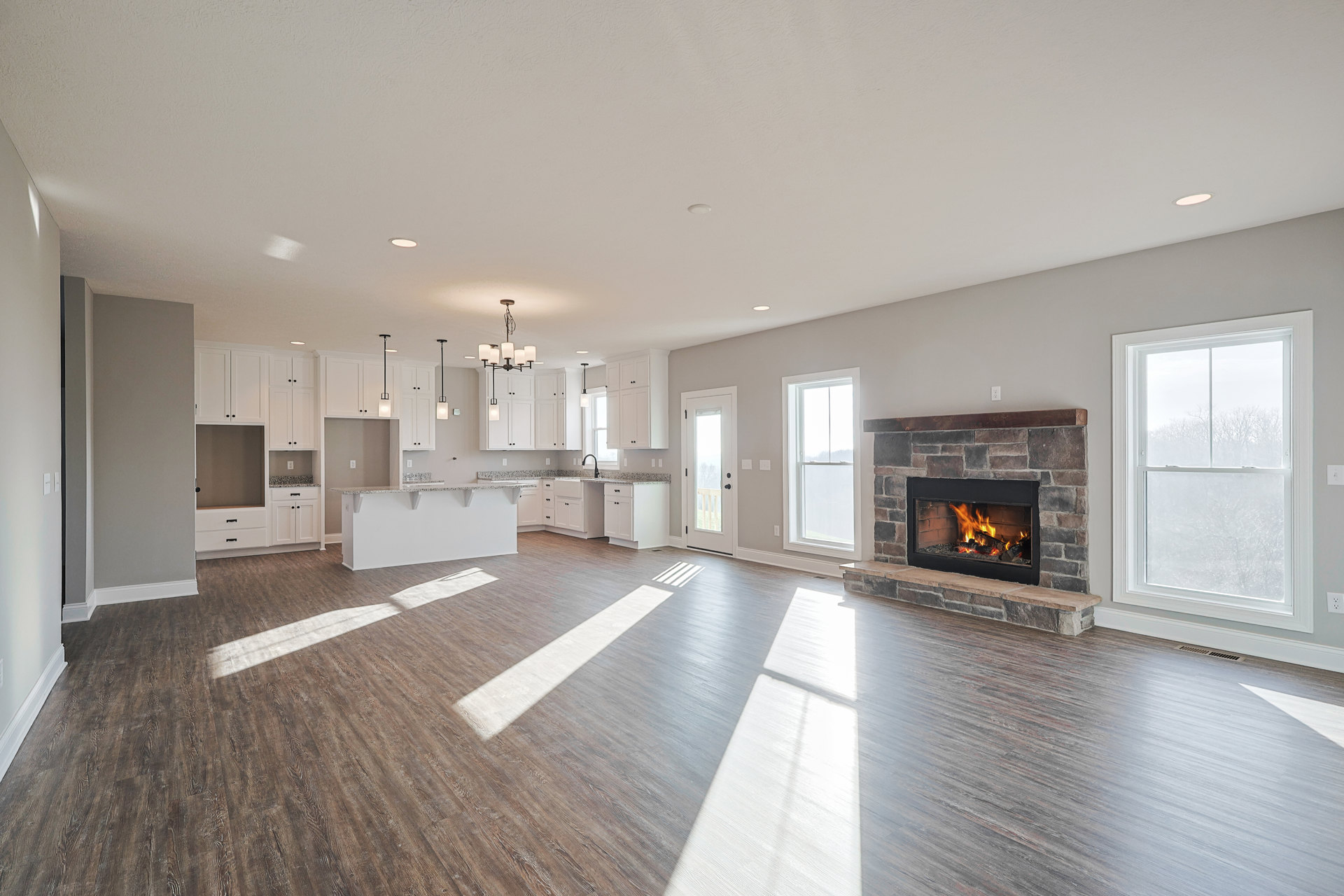 Spacious living room with wood flooring, tile fireplace with burning fire, large white-framed window overlooking trees, and glass door with railing