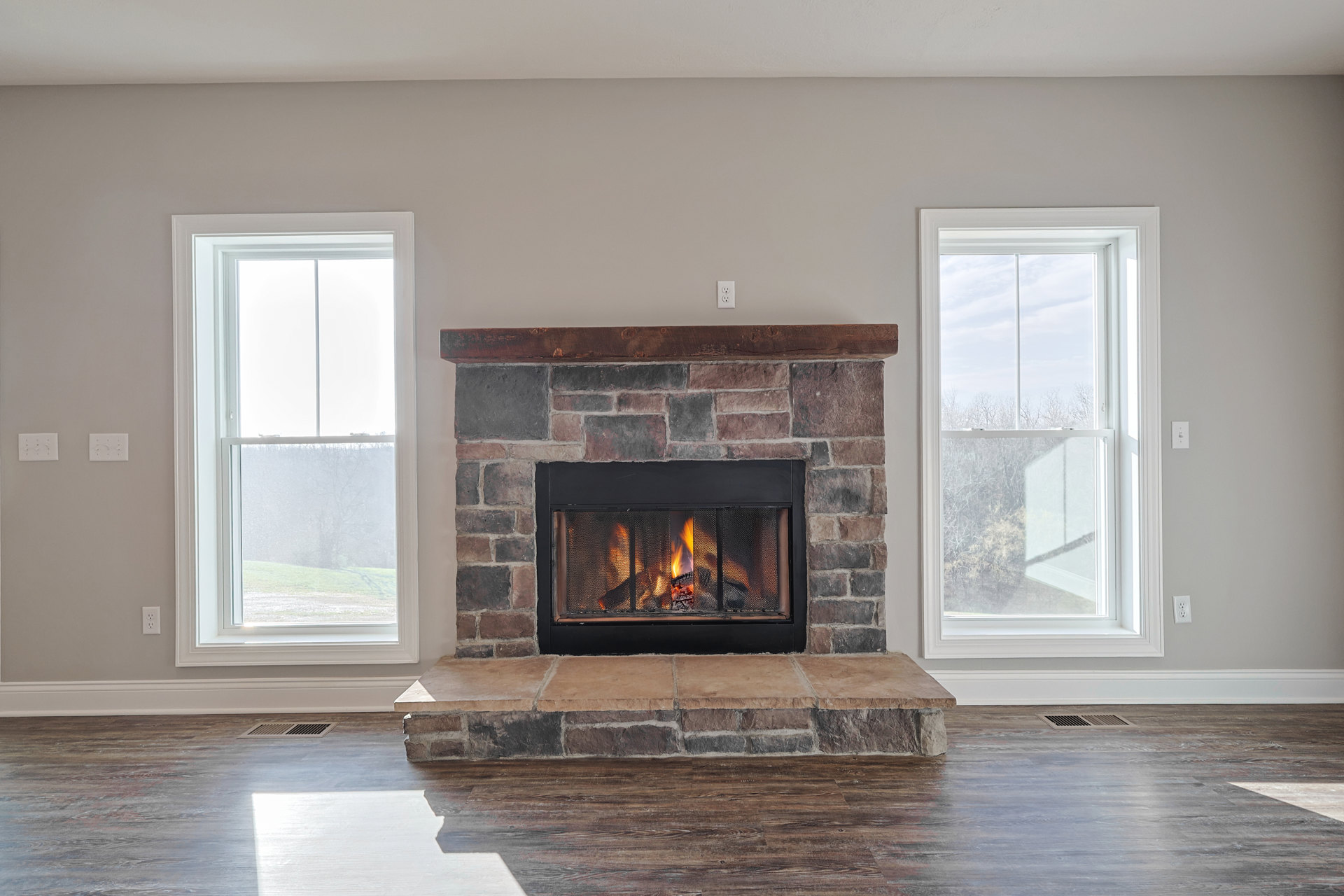 Brick fireplace with black trim, wood-burning fire, wood flooring, large window framing leafy trees, neutral walls, cozy living room ambiance