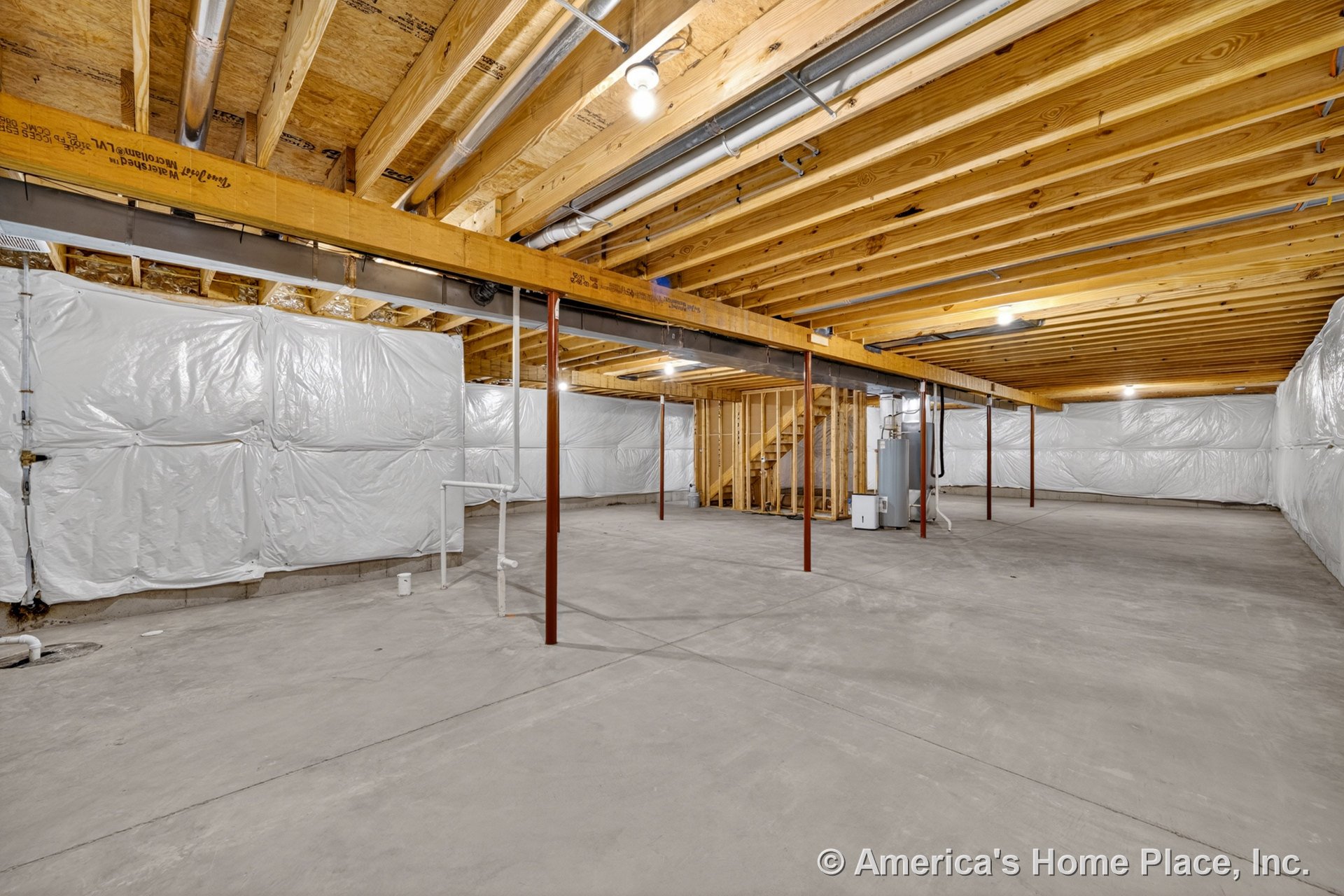Unfinished basement with exposed wood ceiling joists, concrete slab flooring, insulated plastic-wrapped walls, metal support columns, visible plumbing pipes, and a utility area