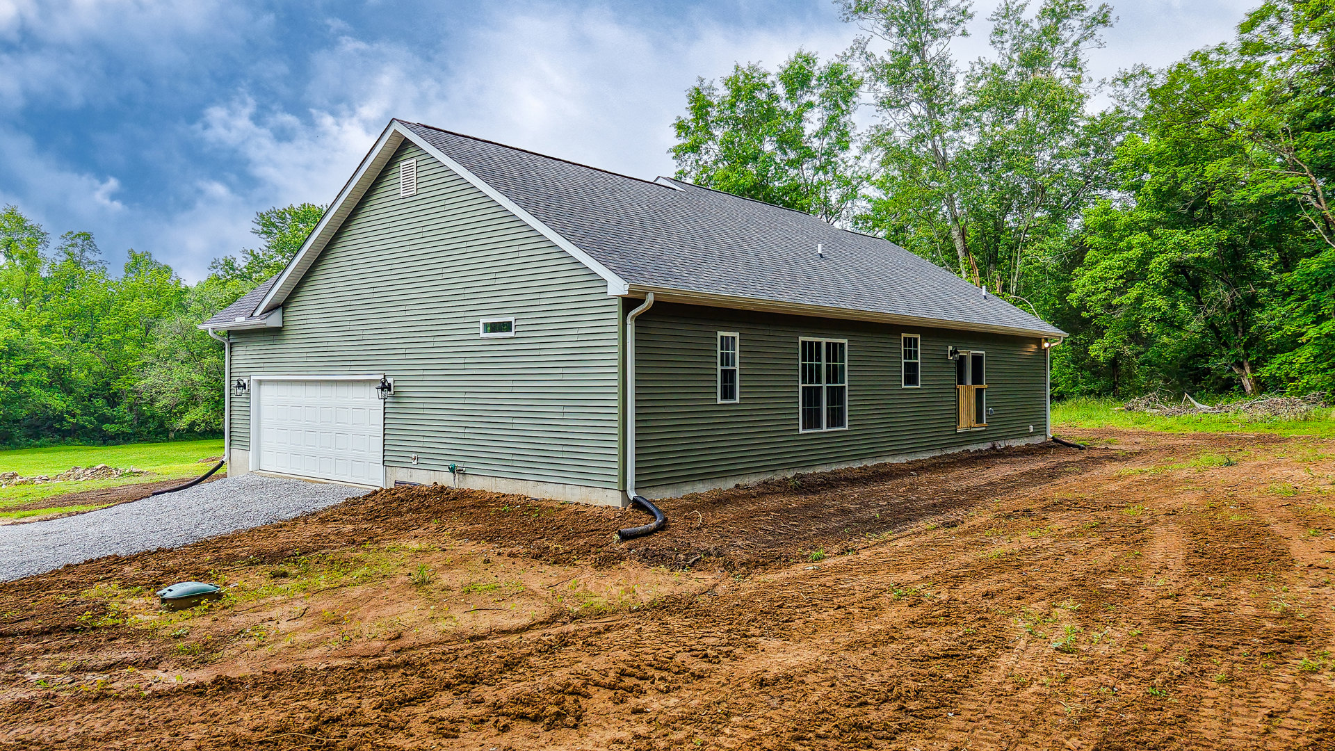 White garage door beneath green roof, white-framed windows, wooden slat accent, gutter system, exterior pipe at house corner, surrounded by grass and trees under cloudy sky