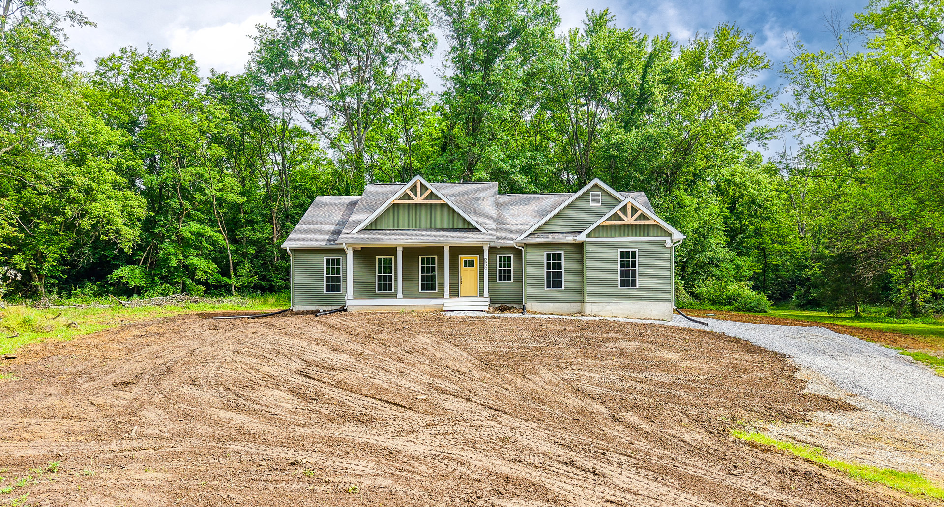 White cottage-style house with yellow front door and window, surrounded by a plowed dirt field, leafy tree, and grassy lot under a blue sky