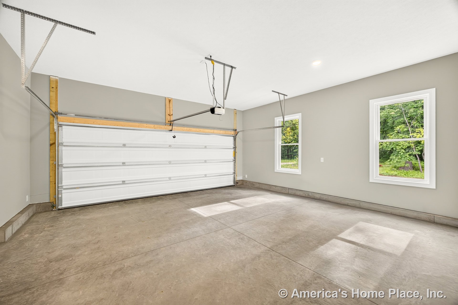 Spacious garage with large white sectional overhead door, smooth concrete floor, two double-hung windows with white trim, ceiling-mounted garage door opener, neutral painted walls